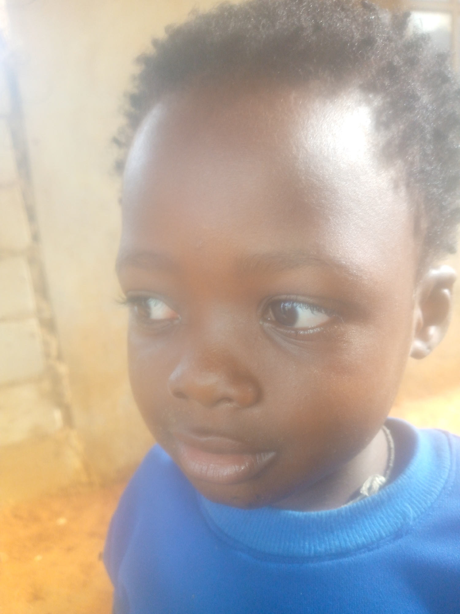 A close-up portrait features a young child, likely a toddler or preschooler, with dark skin and short, dark, curly hair. The child is looking slightly to the right with a neutral, perhaps curious, expression. The image is brightly lit, with strong light coming from the left, creating a prominent highlight on the child's forehead and left cheek, and a soft, hazy glow over the scene.
The child is wearing a vibrant blue crew-neck long-sleeved shirt or sweatshirt. A light-colored necklace or cord is visible around the child's neck, partially resting inside the shirt collar.
The background is out of focus, dominated by warm, earthy tones of light brown and yellow-orange. Subtle rectangular patterns on the far left suggest a textured wall or building structure. The scene appears to be outdoors, under direct sunlight, indicating it is daytime. There is no specific activity taking place other than the child's observed gaze. No text is visible in the image.