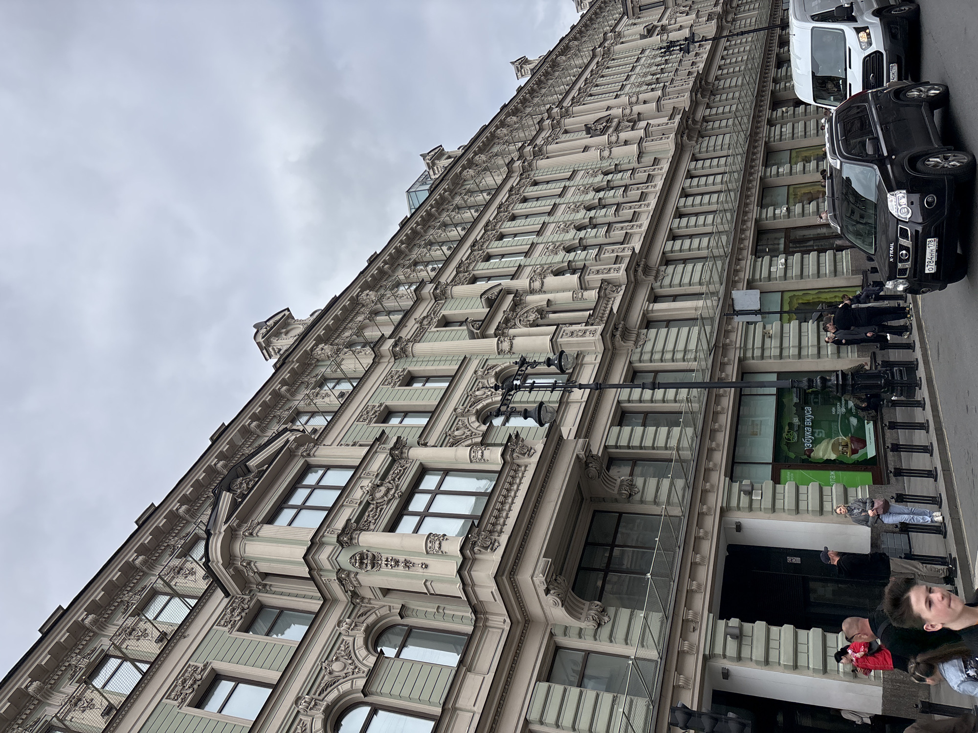 The image shows a tall, ornate building in St. Petersburg, Russia. The building has multiple floors with numerous windows and intricate architectural detailing. A street is visible in the foreground, with pedestrians and parked cars. A white van and black SUV are parked next to the building. A group of people stand near the entrance of the building. The sky is overcast. The location is identifiable by the architecture, which is characteristic of St. Petersburg.
