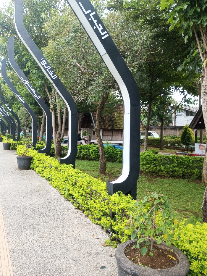 The scene depicts a landscaped pathway in Daringbadi, India. In the immediate foreground, a grey concrete path with a faint brown tactile strip on its left edge extends from the bottom of the frame towards the background. A dark grey cylindrical planter containing a small, sparsely leafed plant rests on the path in the bottom right corner. Parallel to the path, a manicured hedge of vibrant light green shrubbery forms a border.
Behind the hedge, a series of identical, tall, black, curved, arch-like structures with white inner edges stand upright, receding into the distance. Each structure prominently displays white Arabic script vertically. Visible scripts include "الجمال" (Al-Jamal), "الخالق" (Al-Khaliq), "القدوس" (Al-Quddus), "القهار" (Al-Qahhar), and "الرزاق" (Ar-Razzaq), suggesting names of Allah.
Beyond these structures lies a green lawn interspersed with numerous trees of varying sizes and green foliage. In the background, several buildings are discernible: a rustic structure with a dark, possibly tiled, roof and woven-panel walls is situated to the left. Further right, a lighter-colored building with a dark roof is visible behind the trees. A white van is partially visible between the trees. To the far right in the background, a small structure with a dark roof and a conical evergreen shrub in front of it are present, along with a partially visible red and white sign. The environment is outdoors, with lush vegetation under overcast lighting conditions. No identifiable individuals or specific actions are discernible.