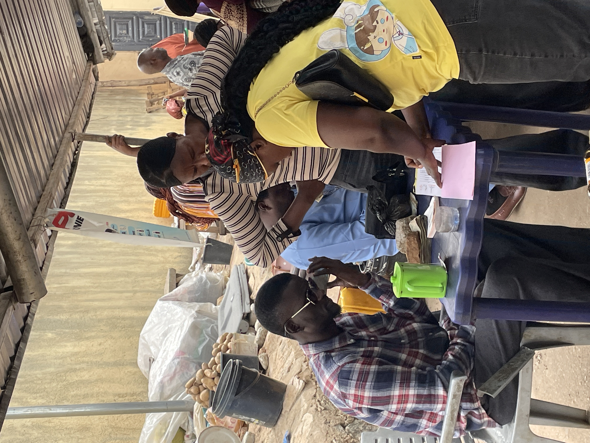 A group of people are gathered around a table outdoors, likely at a polling station in Bukuru, Nigeria. One man, wearing a plaid shirt and glasses, is seated at the table and appears to be interacting with a cell phone. Another person in a light blue shirt is adjacent to him, also looking towards the table. Two women, one in a striped shirt and another wearing a yellow shirt, are standing by the table looking over documents that have been placed in front of them. Additional individuals are visible in the background. Several items are on the table including a pink piece of paper and a green cup. A pile of food items, including potatoes, is visible beside a black bucket on the ground near the table. The area is partly covered by a metal awning.