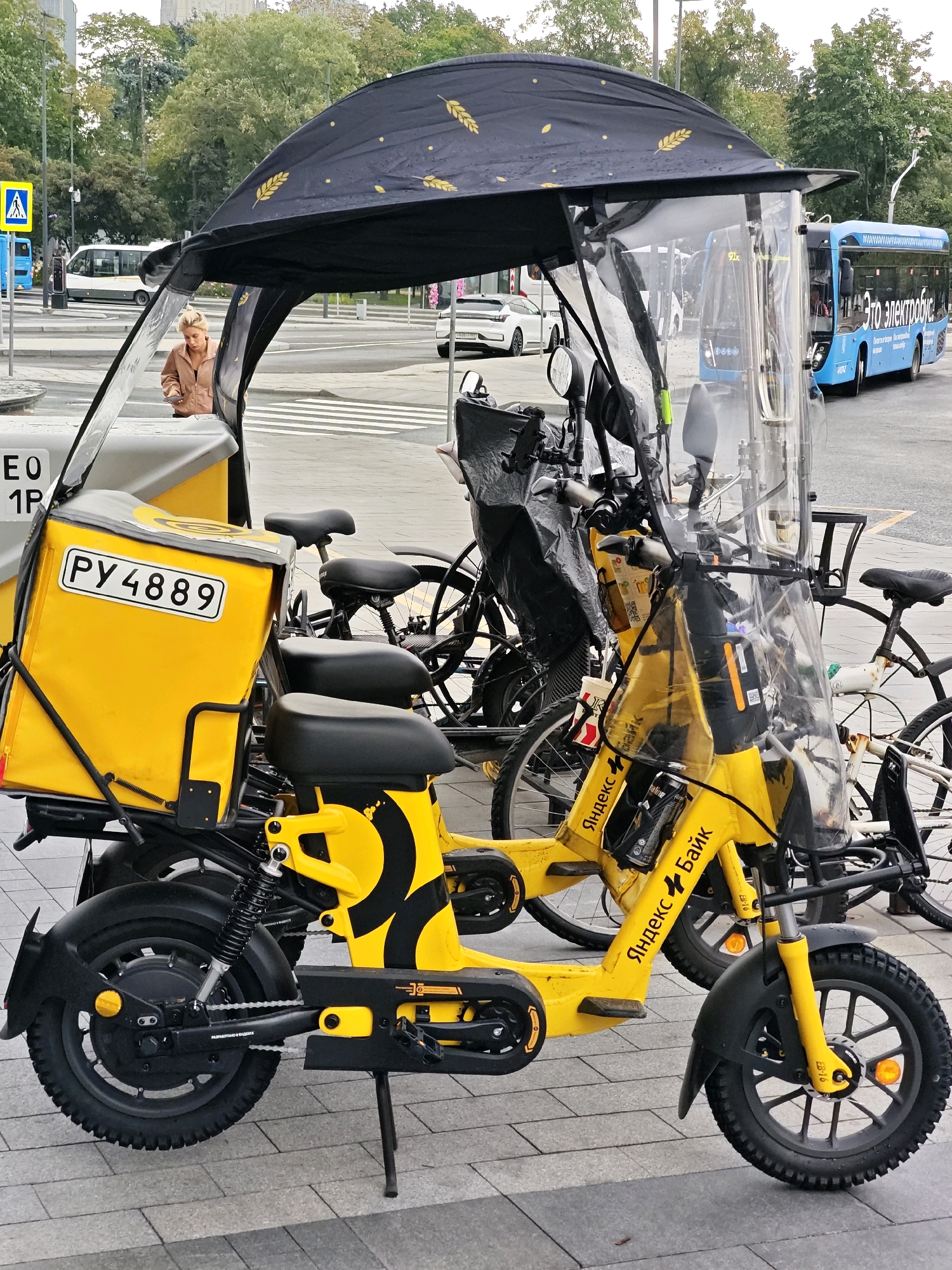A yellow electric scooter with a large yellow delivery box on the back is parked on a paved area. The scooter has a clear plastic shield attached to its front, with a black canopy overhead. A person wearing a tan jacket is standing behind the scooter, looking down at something in their hands. In the background, there is a blue electric bus with Russian text on its side, a white bus, and a white car. Trees and buildings are visible in the distance. The scene appears to be a public street or transport hub in Moscow, Russia.