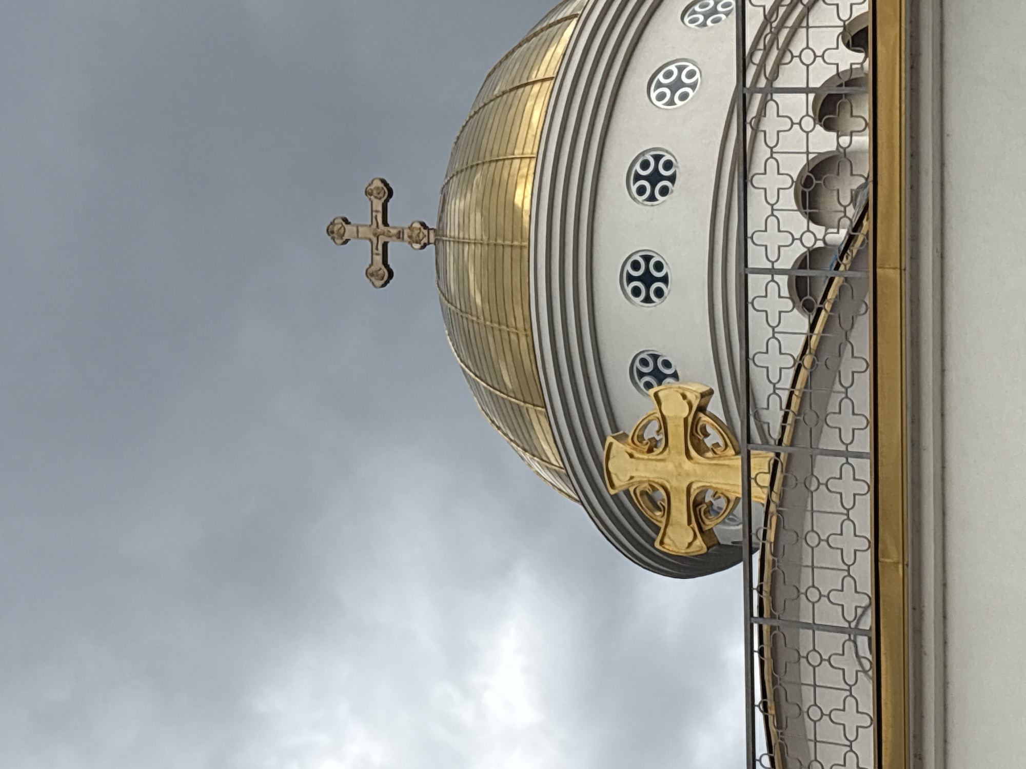 The image captures a detail of the Church of St. John Vladimir in Bar, Montenegro, specifically focusing on the golden dome adorned with crosses against a cloudy sky. The dome is ribbed with gold and features a row of small, round windows along its base. A golden cross is affixed to the side of the dome, with another smaller, more ornate cross at the top. To the right of the dome is a light-colored architectural element, potentially part of the church facade, featuring a decorative metallic grid with cross-shaped motifs. The backdrop consists of an overcast sky, providing a muted contrast to the golden accents of the church.