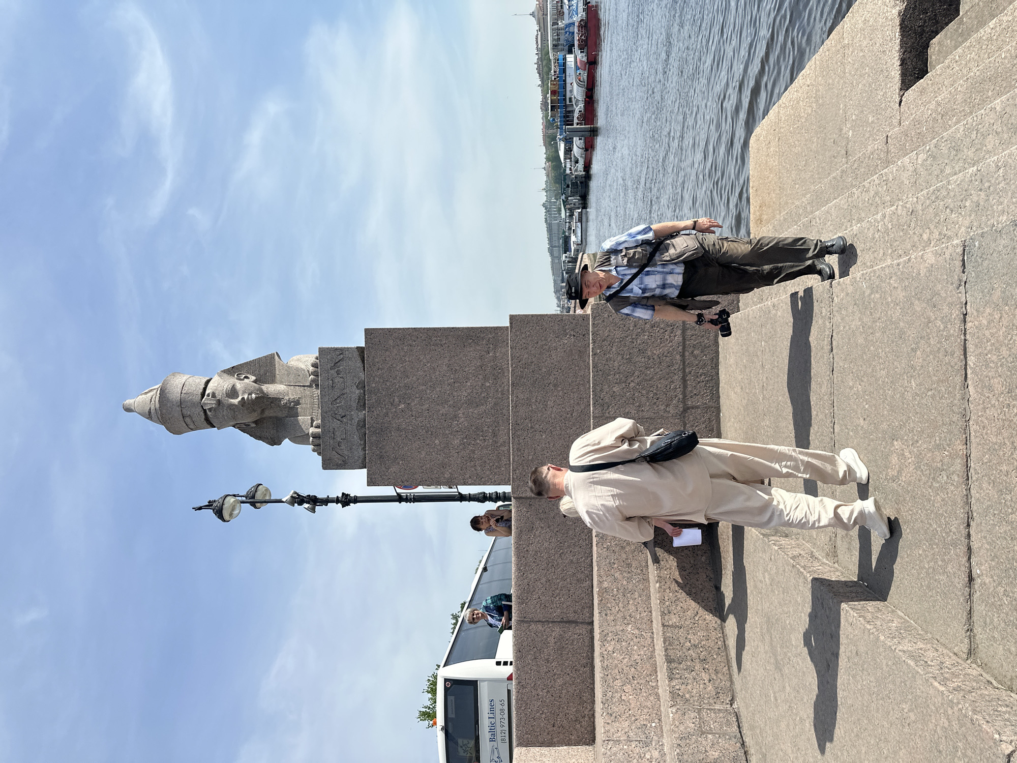 The image depicts a scene at the Egyptian Sphinxes on the Universitetskaya Embankment in Saint Petersburg, Russia. A gray granite sphinx statue sits atop a granite pedestal, facing towards the sky. A lamppost is adjacent to the statue. Two men are on the steps: one wearing a plaid shirt, dark pants, and a hat, with a camera in hand, and another wearing a light-colored suit with a black bag. A bus with "Baltic Lines" visible on the side, is parked nearby. In the background, the Neva River with various boats and buildings can be seen under a blue sky.