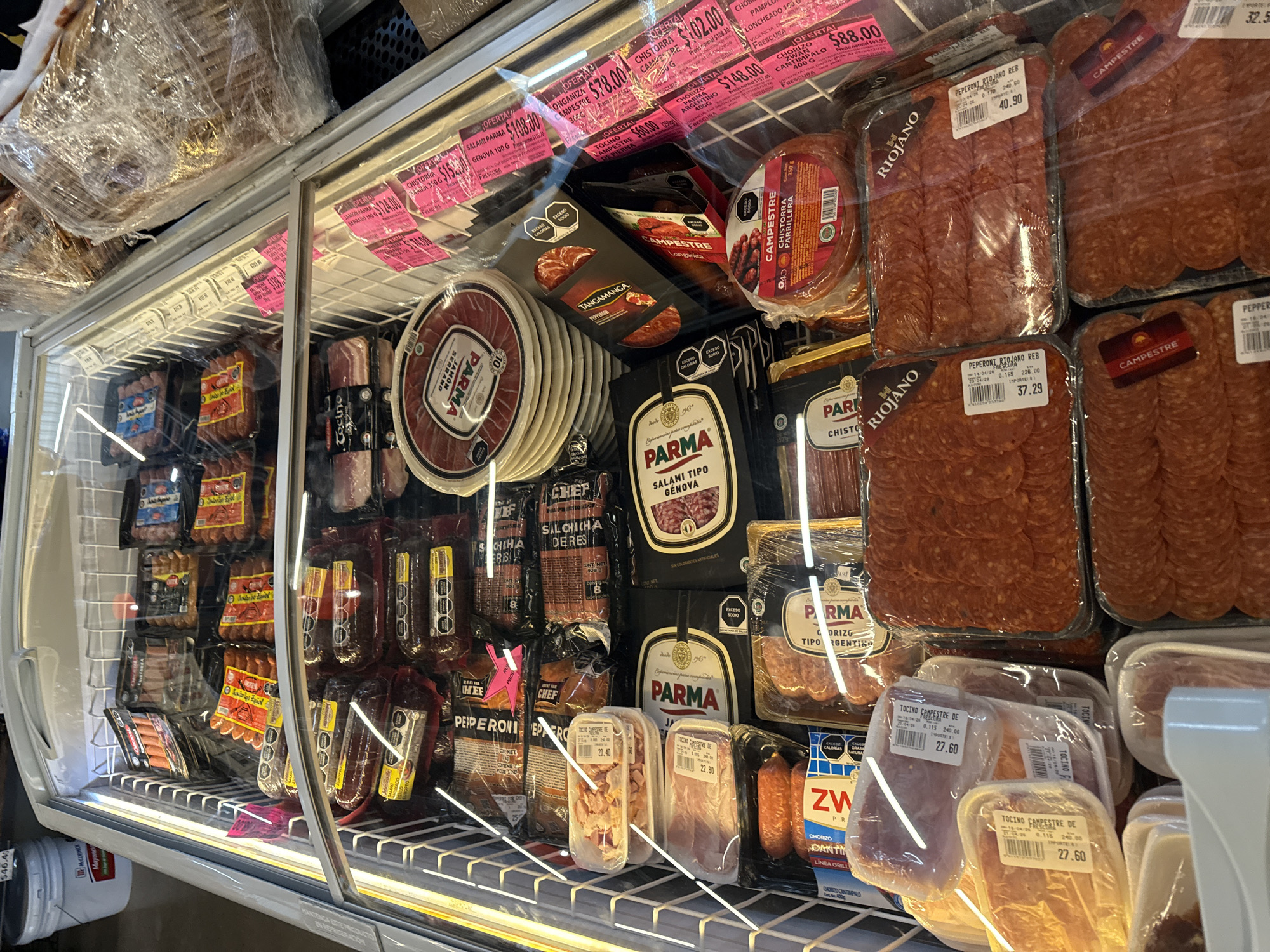 This image captures a refrigerated display case filled with a wide variety of packaged meat products, likely in a grocery store or deli in Puebla, Mexico. No people are visible.
The shelves are packed with numerous types of cold cuts, sausages, and cured meats. Dominant brands include "Parma," offering products like "Jamon Serrano," "Salami Tipo Genova," and "Chorizo Argentino." "Campestre" brand is also prominent, with "Chistorra Campestre," "Longaniza," and "Tocino Campestre de Pierna" (bacon). Other visible brands include "Riojano" (featuring pepperoni), "Chef," "Tangamanga" (with pepperoni), and "Zwan" (chorizo cantimpalo).
Many products are marked with vibrant pink "OFERTA!" (offer) labels displaying prices in Mexican Pesos, such as $108.00 for Parma Salami, $78.00 for Campestre Longaniza, and $88.00 for Zwan Chorizo Cantimpalo. Individual product labels also show specific weights and prices, for instance, "Peperoni Riojano Reb" for $40.90 and $37.29, and "Tocino Campestre de" for $27.60.
Additional text on packaging includes nutritional warnings like "EXCESO CALORÍAS" (excess calories) and "EXCESO SODIO" (excess sodium), as well as "SIN COLORANTES ARTIFICIALES" (no artificial colors), and instructions like "MANTENGA ESTE PRODUCTO EN REFRIGERACIÓN" (keep this product refrigerated). A "McCormick Mayone" label is partially visible on a container outside the case. The items are neatly arranged, showcasing the array of options available to customers.