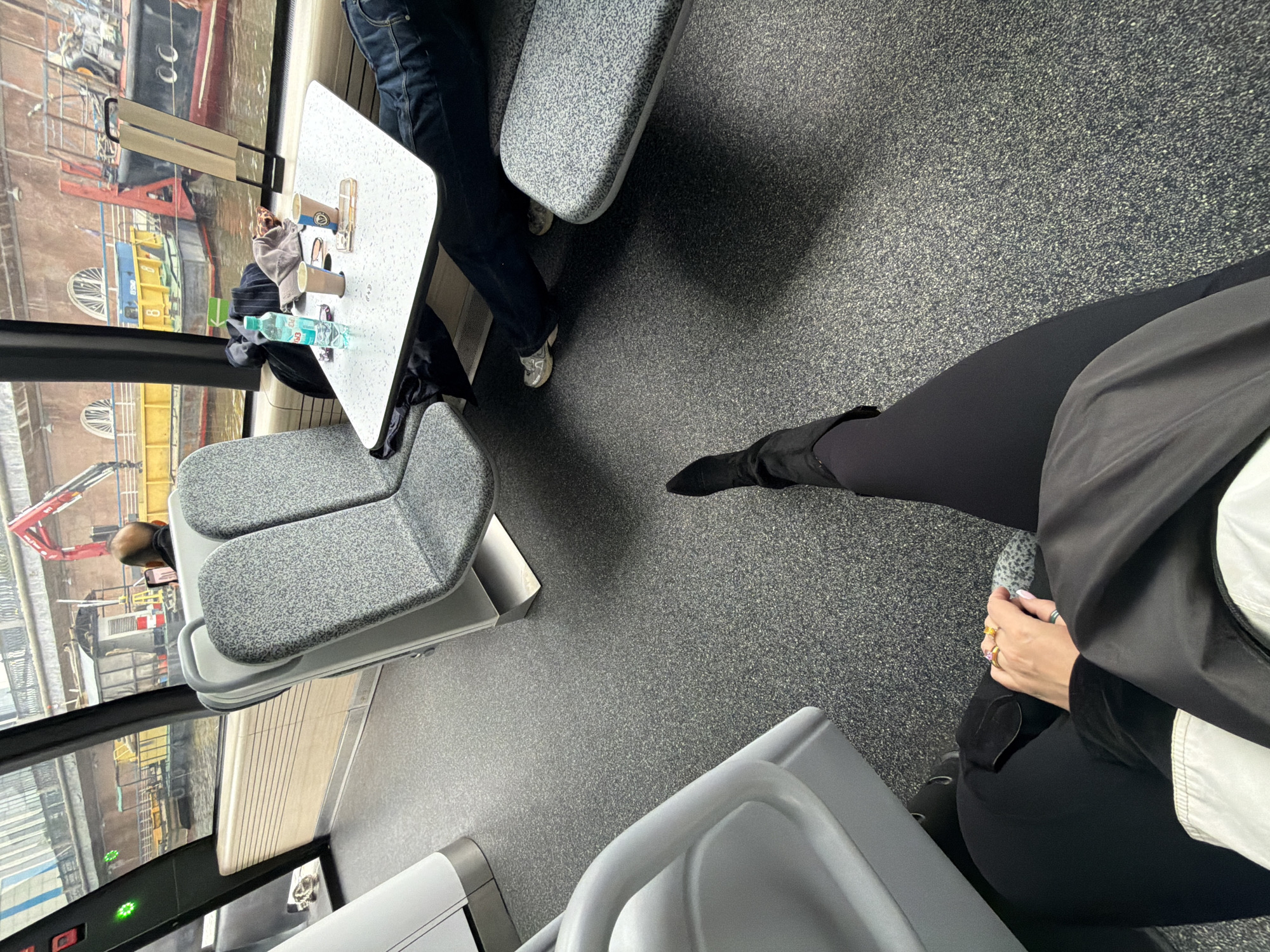 The image depicts an interior shot of a public transport vehicle, possibly a train or bus, in Moscow, Russia. The view is from above, angled downward, capturing seats, tables, and flooring, as well as partial views through the window.
The seating arrangement is booth-style, with two tables present.
One person is visible on the right side of the frame, wearing black pants, a black jacket, a white undershirt, and black boots. The individual's hand, adorned with multiple rings, is visible.
Another person is partly visible at the top of the frame. This person wears denim jeans and grey sneakers.
Cups and bottles are on the table.
Outside the window, there are buildings, cranes, and other industrial elements visible. The environment suggests a port or dock area.
The floor is grey with a grainy pattern.
