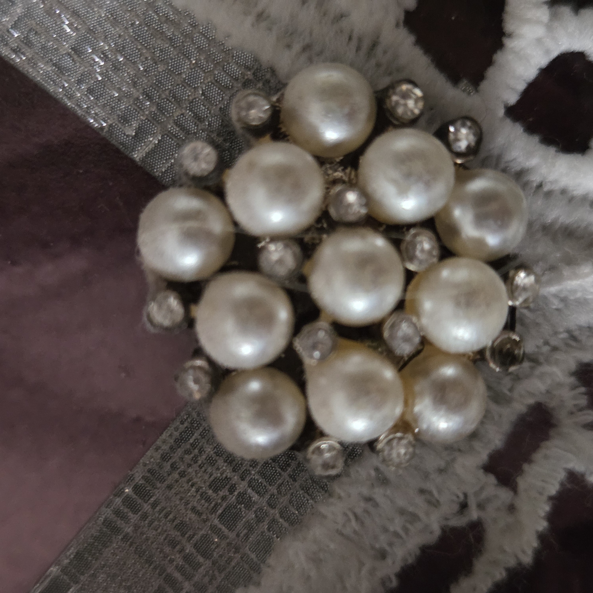 The image displays a close-up of a decorative embellishment, likely a brooch or part of an accessory. The central object is a circular cluster of numerous off-white to cream-colored pearls, varying slightly in size, arranged closely together. Interspersed between the pearls are smaller, clear, faceted rhinestones that add a sparkling accent. The pearls exhibit a soft luster, reflecting light gently.
The embellishment rests on a multi-textured surface. To the left, a dark purplish-brown material, possibly velvet or a smooth fabric, provides a muted background. Below and to the left, a silvery-grey fabric or material with a distinct, textured pattern of small, repeating squares or diamonds is visible. This material has a subtle sheen, catching reflections. To the right and upper right, a fluffy, light-colored material, appearing to be faux fur or a soft, textured fabric, is present, featuring irregular dark shapes within its pile.
The lighting is soft, highlighting the pearlescent quality of the spheres and the sparkle of the rhinestones. No specific activity is depicted, nor is any text visible. The image captures the intricate details and textures of the ornamental piece and its immediate surroundings.