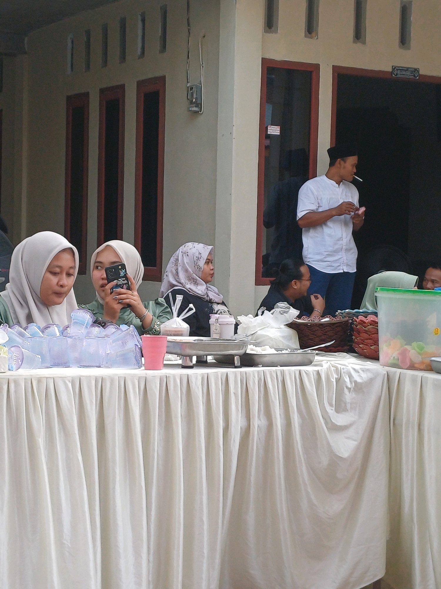 In Dumai City, Indonesia, at a casual gathering, a man in a white shirt and black peci stands near a doorway, smoking a cigarette and holding a small pink object. In the foreground, three women wearing hijabs are seated behind a table covered with a white cloth. The woman on the left is looking down, while the middle woman is holding a smartphone with a "Stitch" character case and taking a selfie or photograph. The woman on the right is looking to the side. On the table, there are stacks of clear plastic cups, a pink cup, food containers, and a basket. The background shows a building with beige walls and dark-framed windows.