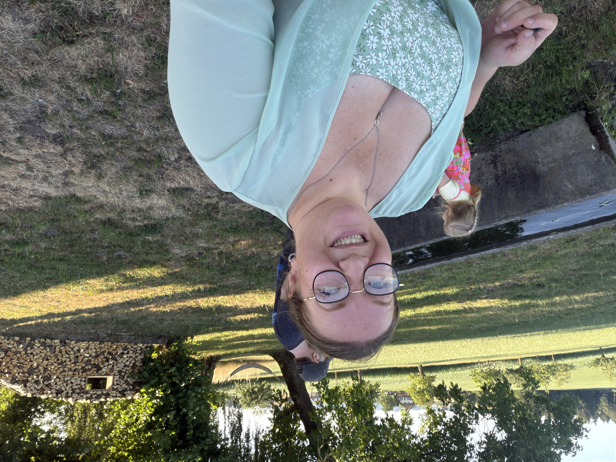 A woman is taking a selfie outdoors. She is smiling and wearing glasses and a green and white floral dress with a light green sheer overlay. In the background, a young child in a pink dress stands near a concrete trough filled with water. Further back, a man in a dark shirt and a green cap is visible, along with a hammock strung between two trees. To the right, a stack of firewood with a small window-like opening is visible. The scene appears to be in a rural area, likely Ufhusen, Switzerland, with a field and trees in the distance. The sun is low, casting long shadows.