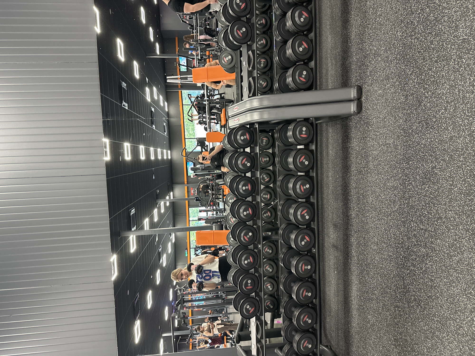 The image shows the interior of a gym, potentially located in Plovdiv, Bulgaria. A person is performing bicep curls with dumbbells, facing towards a mirrored wall. Racks of dumbbells line the side of the frame. Other gym equipment is visible in the background, and several other people can be seen exercising in the gym. The floor appears to be a textured, dark grey material. The gym has a black ceiling with rectangular lights.