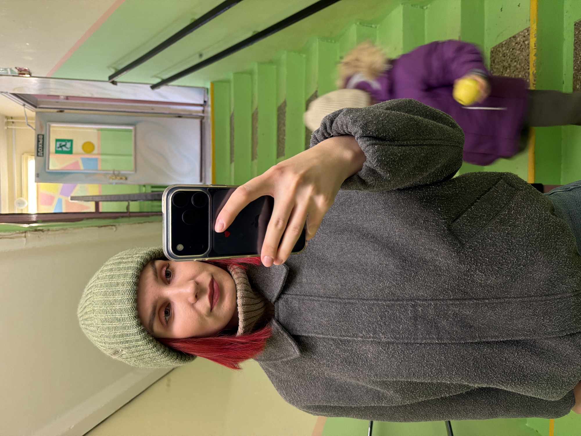 A young woman is taking a selfie in an indoor stairwell, holding a black smartphone with her left hand. She has red hair and is wearing a light green knit beanie, a high-neck beige/tan ribbed sweater, and a textured dark gray coat. Her expression is neutral to slightly smiling.
In the blurred background, a child or smaller person is visible, walking down bright green stairs. They are wearing a purple coat with a light-colored, possibly fur-trimmed, hood and holding a yellow object, possibly a ball or toy.
The setting is a brightly lit staircase with prominent green steps and a black handrail running alongside. The walls are light-colored. On a wall in the mid-ground, a reflective surface (likely a mirror or framed glass) shows a reflection of a colorful, abstract wall decoration featuring geometric shapes in yellow, green, light blue, and pink. A green fire exit sign with a running person pictogram and the Russian word "ВЫХОД" (EXIT) is clearly visible on this reflective surface. The overall atmosphere is that of a public or institutional building in Chaikovsky, Russia.
