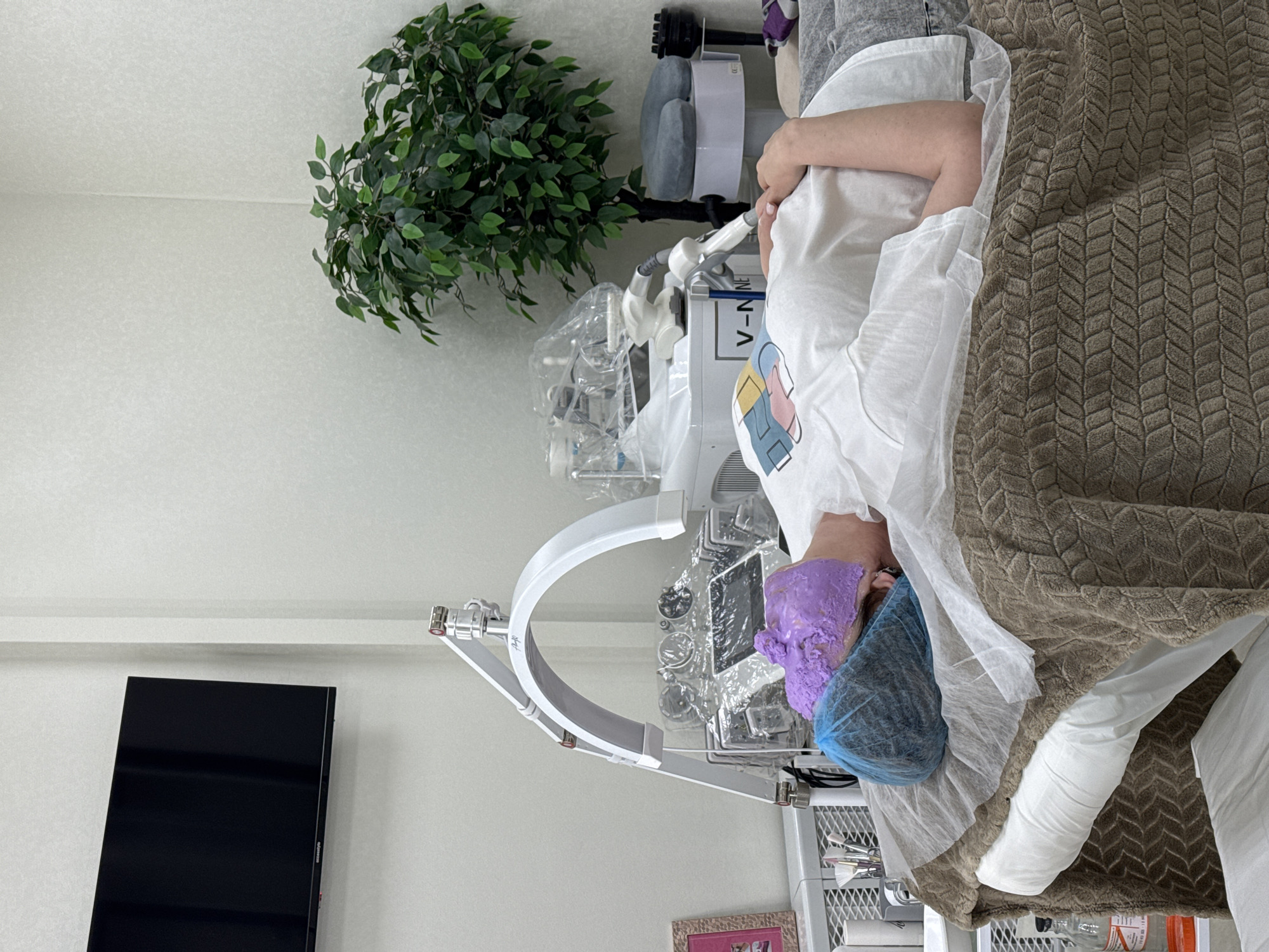 A person wearing a surgical cap and a purple facial mask is lying on a treatment bed covered with a brown patterned blanket and a white sheet. The person is wearing a white t-shirt with a colorful design. An articulated white arm extends over the person's face, likely part of a cosmetic treatment machine. To the left of the treatment bed, there is a black television mounted on the wall, a white storage unit with assorted cosmetic items, and a framed picture on top. Behind the bed, there is a white machine with the letters "V-N" visible. A potted green plant sits in the corner of the room. The location is Unknown, Unknown.