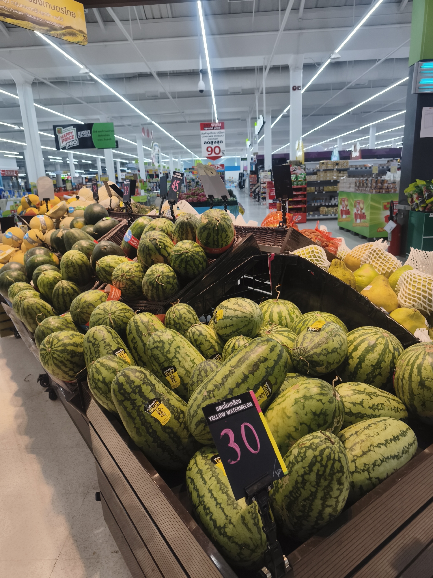 The image captures a display of various fruits, particularly watermelons, in what appears to be a grocery store located in Baan Bo Phut, Thailand. The primary focus is on a large assortment of light green striped watermelons, some labeled as "YELLOW WATERMELON" and priced at "30". A separate sign indicates a discount of up to "90%". Other fruits such as melons and netted yellow fruits are visible in the background. The scene suggests a typical produce section within a well-lit supermarket environment.