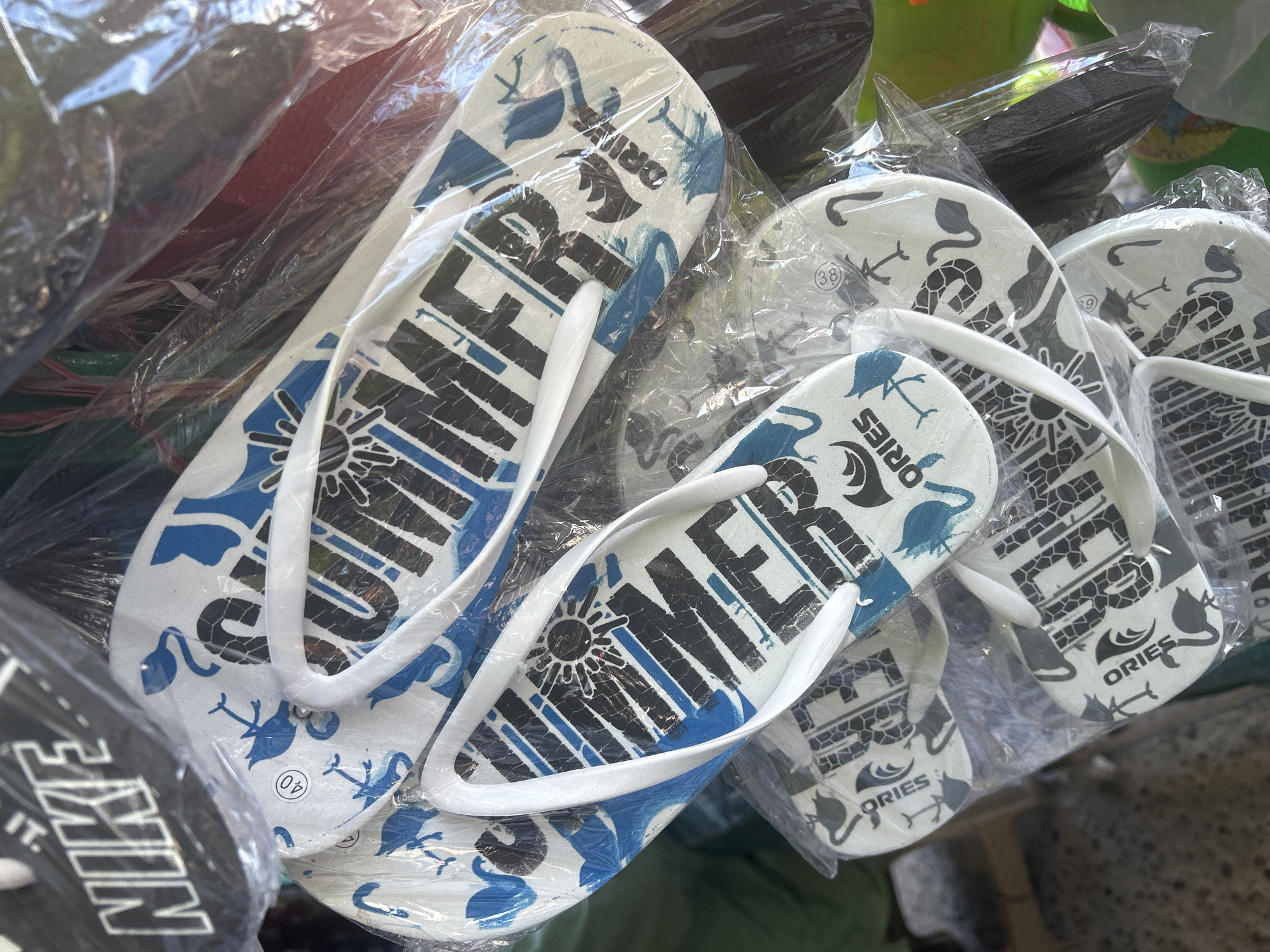 The image displays a close-up of several pairs of flip-flops packaged in plastic, likely at a market stall in Bira, Indonesia. The flip-flops have white soles with blue and black designs. Some soles feature the word "SUMMER" in large, black lettering with a sun graphic above it. Other soles have the brand name "ORIES" and graphics of flamingos and other stylized animals. A black cap with the "NIKE" logo is visible at the top left of the frame. The overall setting appears to be a retail environment, with the items being displayed for sale.