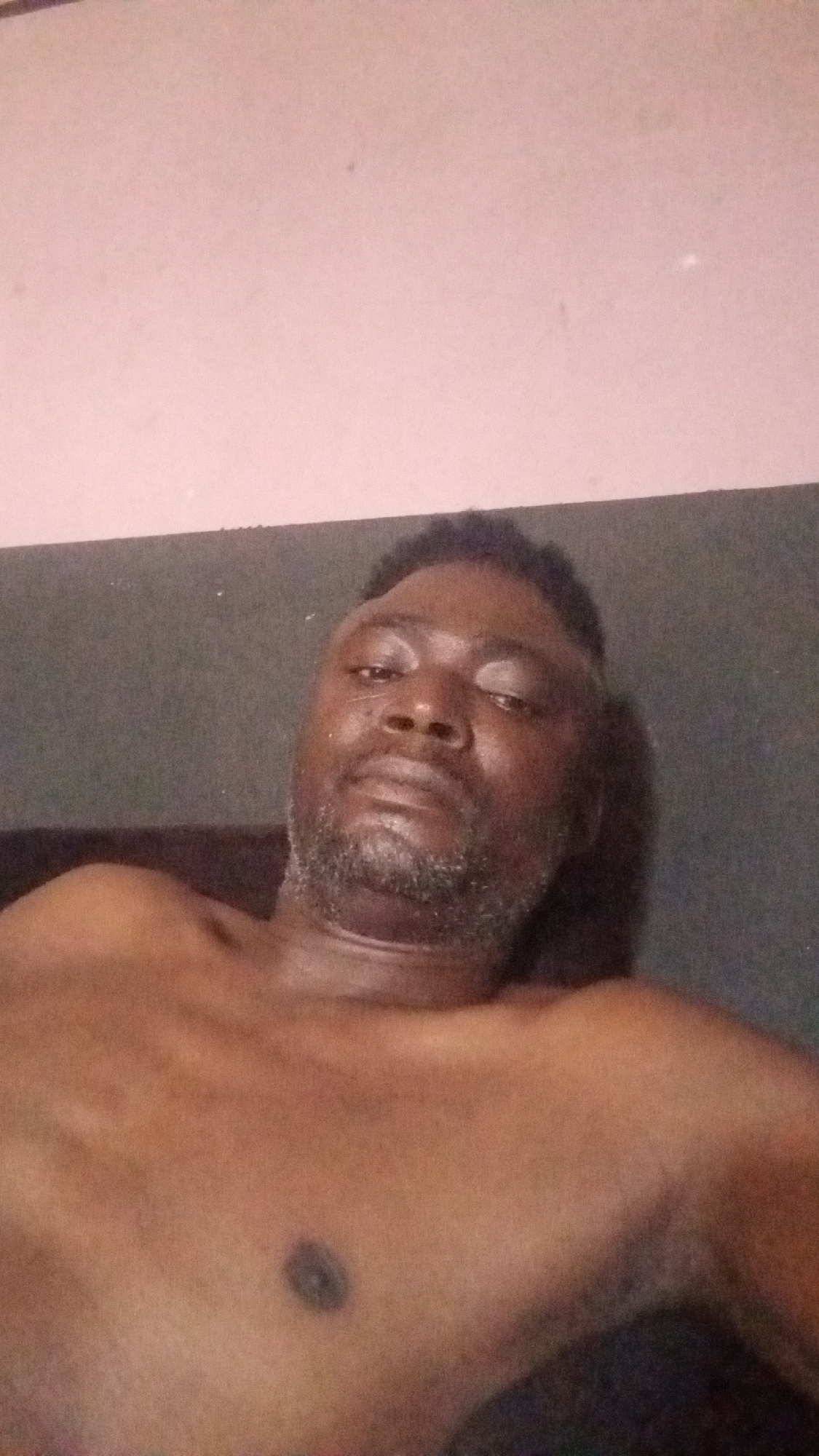 A shirtless middle-aged Black man is lying down. He has a dark complexion, short dark hair, and a beard with some gray hairs. The background shows a light pink wall and a darker gray surface behind his head and shoulders. The location is not discernible from the image.