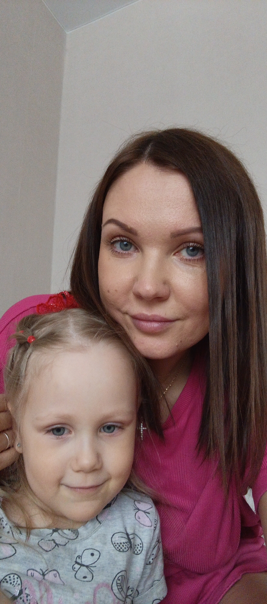 A selfie image captures a woman and a young girl posing together indoors. The woman, with fair skin and dark brown hair parted on the left, is wearing a bright pink sweater. She has light blue eyes and a faint smile. A gold necklace is visible on her neck. The girl, positioned slightly in front of the woman, has light blonde hair with red hair ties, blue eyes, and a slight smile. She wears a gray long-sleeved shirt adorned with butterfly patterns. The background is a plain, light wall. Given the prompt, this photo may have been taken in Chaikovsky, Russia.