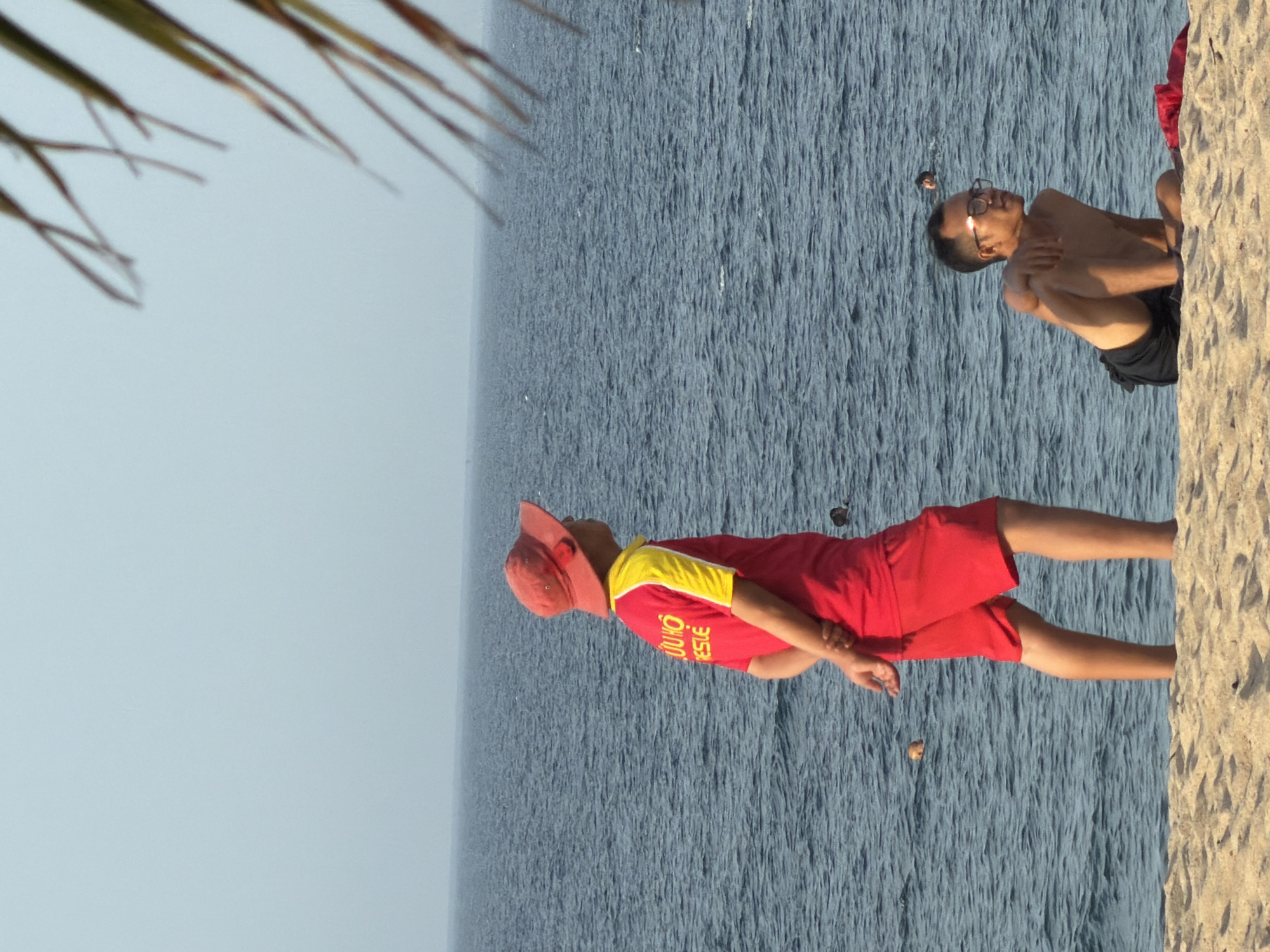 A lifeguard in a red hat and yellow-sleeved shirt stands on a sandy beach, facing right, with hands clasped behind his back. In the background, the blue ocean stretches to the horizon. To the right, a man in black shorts sits on the sand, looking to the right. The scene appears to be a public beach in Nha Trang, Vietnam.