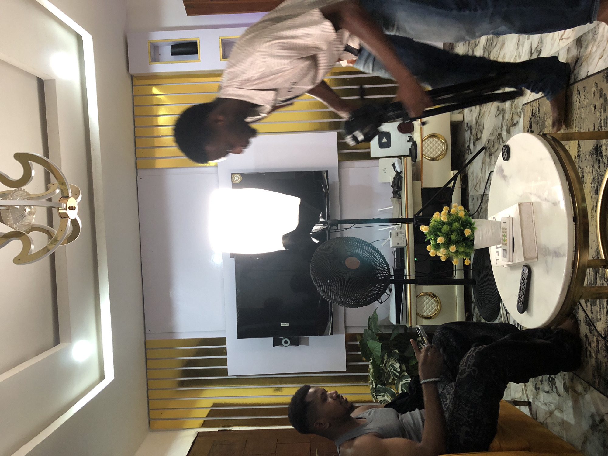 The image captures an indoor scene, likely a living room or studio space, with two men engaged in activities related to content creation. The overall aesthetic is modern, featuring white walls with horizontal gold accents and recessed lighting.
In the foreground, on the right side of the vertically oriented image, a man stands, dressed in a striped button-down shirt and blue jeans. He is focused on adjusting or setting up camera equipment mounted on a black tripod. His face is visible in profile, looking down at the gear. Behind him, a bright softbox light illuminates the area.
Further into the room, a second man is seated on a yellow sofa or lounge chair, wearing a grey tank top and patterned dark pants. He is looking at a smartphone held in his hands, appearing relaxed.
The room is furnished with a white, circular coffee table with gold-colored legs, on which a small vase of yellow artificial flowers, a remote control, and a white box are placed. A black electric fan is positioned near a wall-mounted television screen. Decorative elements include a stylish ceiling light fixture, white shelving with gold trim, and green artificial plants. A white box prominently displaying the "YouTube" logo is visible on a cabinet behind the standing man. The floor features both a marble-like pattern and a dark patterned rug.
The setup, including the camera, tripod, and softbox light, strongly suggests that a video recording or photoshoot is either in progress or being prepared. The room is brightly lit by artificial lights.