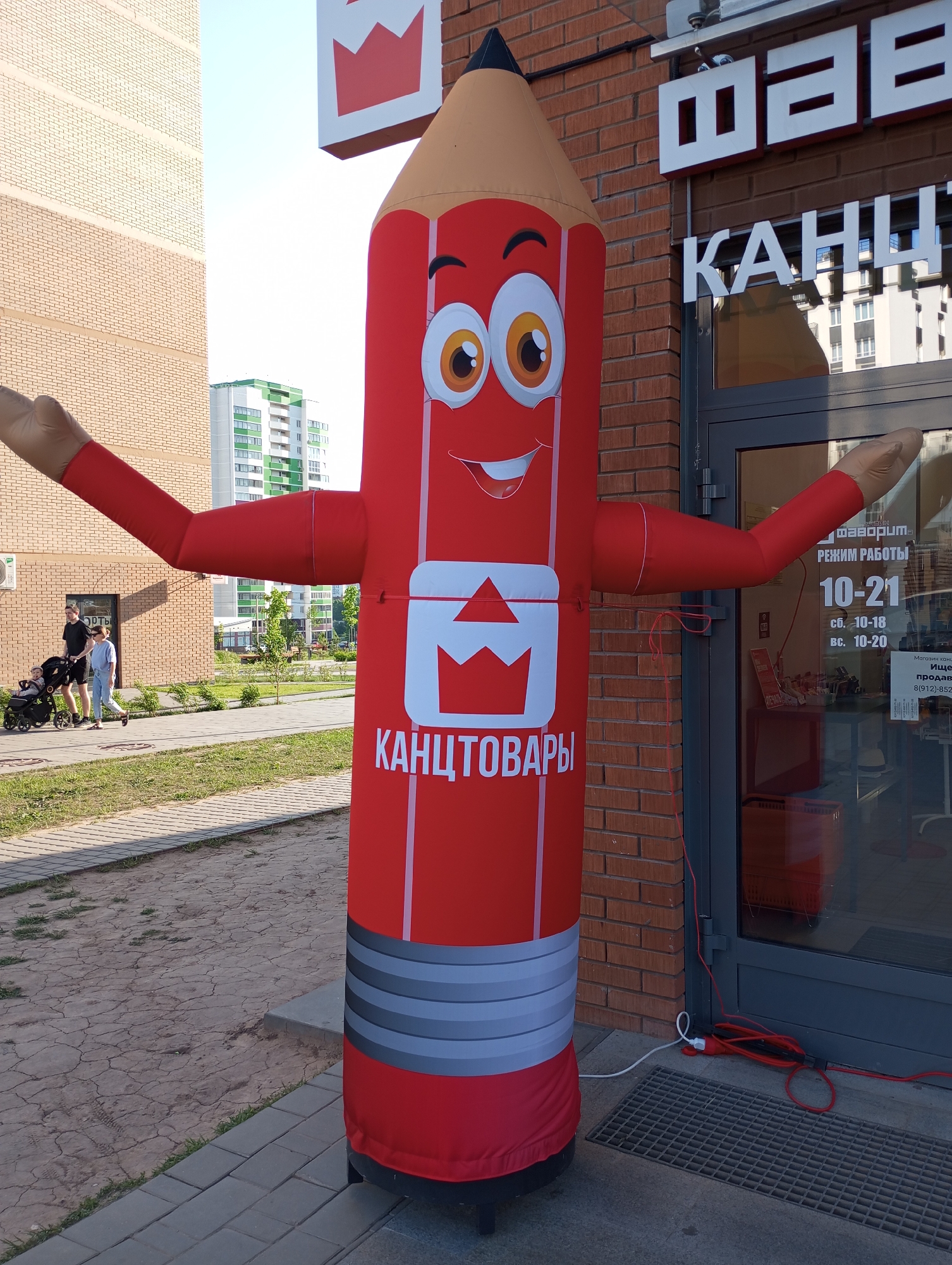 The image shows an inflatable, red pencil-shaped advertising figure with a smiling face and outstretched arms, standing in front of a store in Pervomaysky, Russia. The pencil is decorated with the logo of the "Favorit" (Фаворит) stationery store and the word "канцтовары" (stationery). The store entrance is behind the figure, displaying opening hours. A family is walking on a sidewalk in the background. The environment includes brick buildings, green grass, and paved walkways.