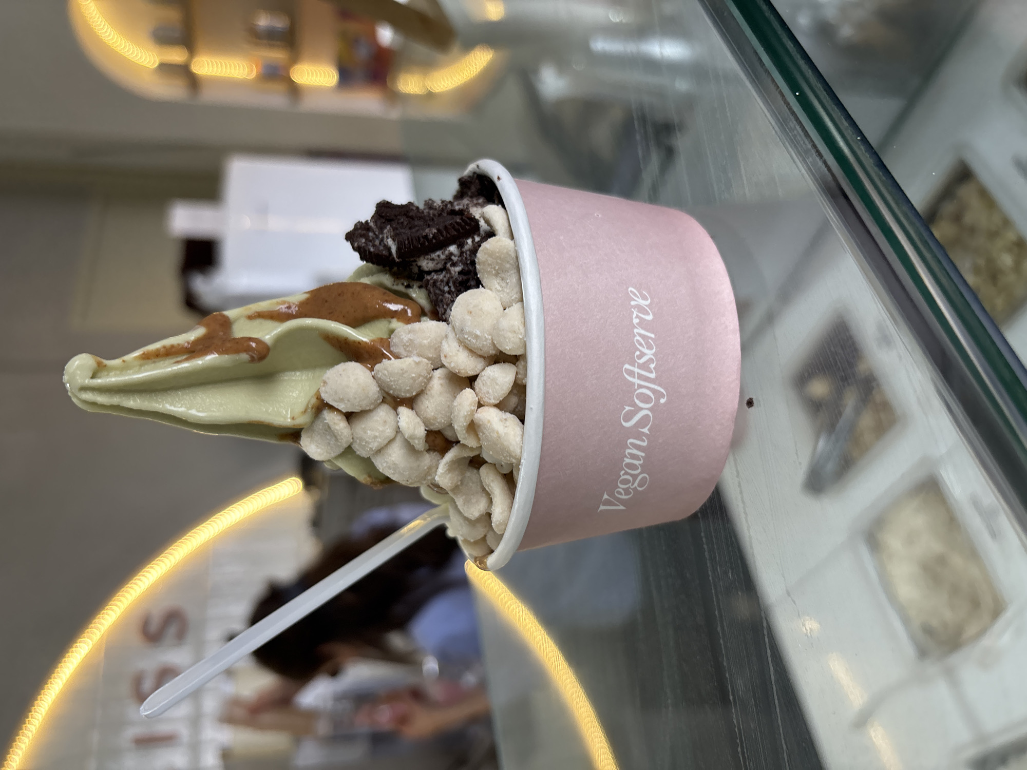 A cup of vegan soft serve ice cream from "Vegan Softserve" is displayed in a clear display case. The soft serve is light green and drizzled with brown sauce. It is topped with white-coated candies and crushed Oreo cookies. A white plastic spoon is inserted into the cup. The background shows blurred lights and other items in the display case. The location of the image is in San Bernardino Tlaxcalancingo, Mexico.