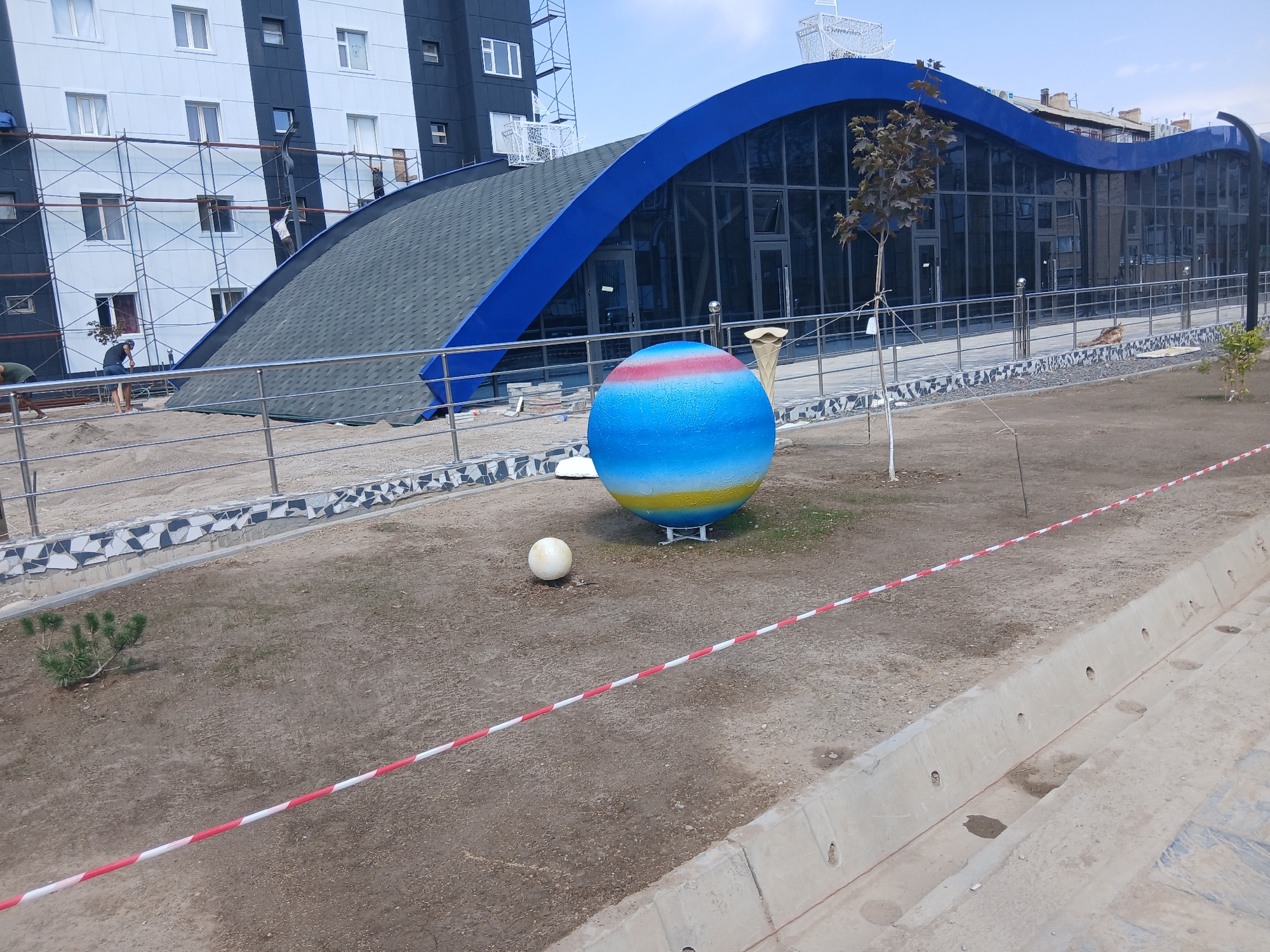 The image depicts a construction site in Talas City, Kyrgyzstan, with a partially completed building featuring a distinctive wave-shaped blue roof and glass facade. Scaffolding surrounds a section of the building, indicating ongoing work. In the foreground, a large, colorful sphere painted with bands of blue, yellow, and red rests on the ground. A smaller white sphere is situated nearby. A red and white striped tape is laid across the dirt ground, marking off an area. A small evergreen tree stands to the left of the large sphere. The overall scene suggests a public space undergoing development or renovation.