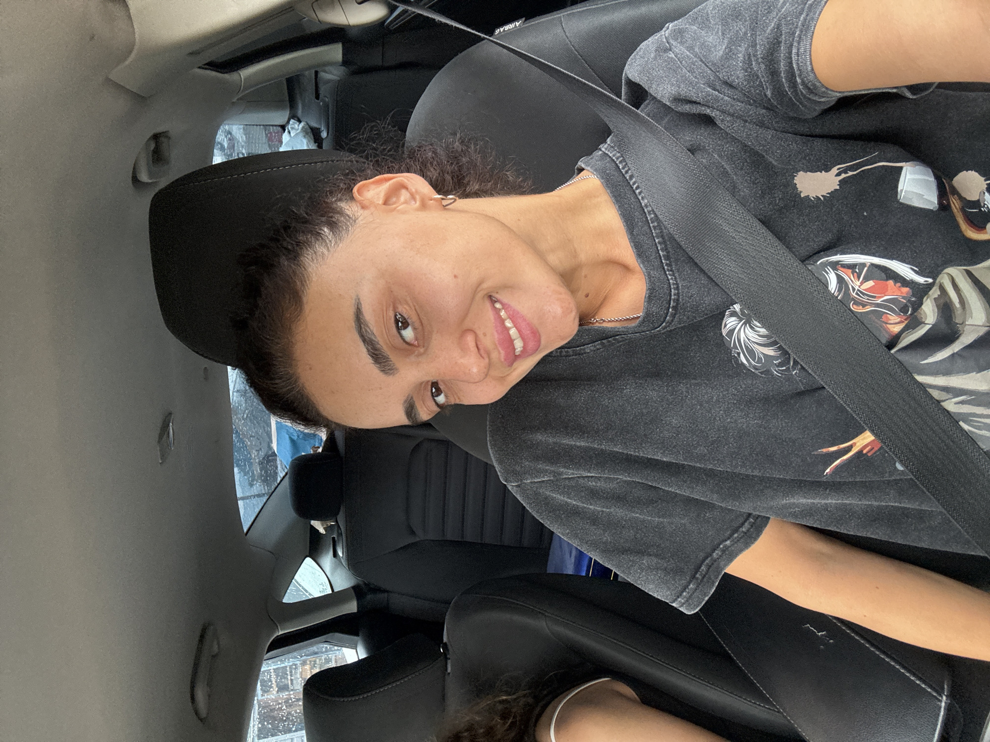A young woman with curly dark hair, wearing a dark grey t-shirt and a seatbelt, smiles at the camera from the passenger seat of a car. She is wearing a silver hoop earring and a delicate silver necklace. The interior of the car is visible, with black seats and the headrest of the driver's seat behind her. The window on the right shows a blurry view of an urban environment, with the suggestion of trees and buildings. The image appears to have been taken in Tbilisi, Georgia.