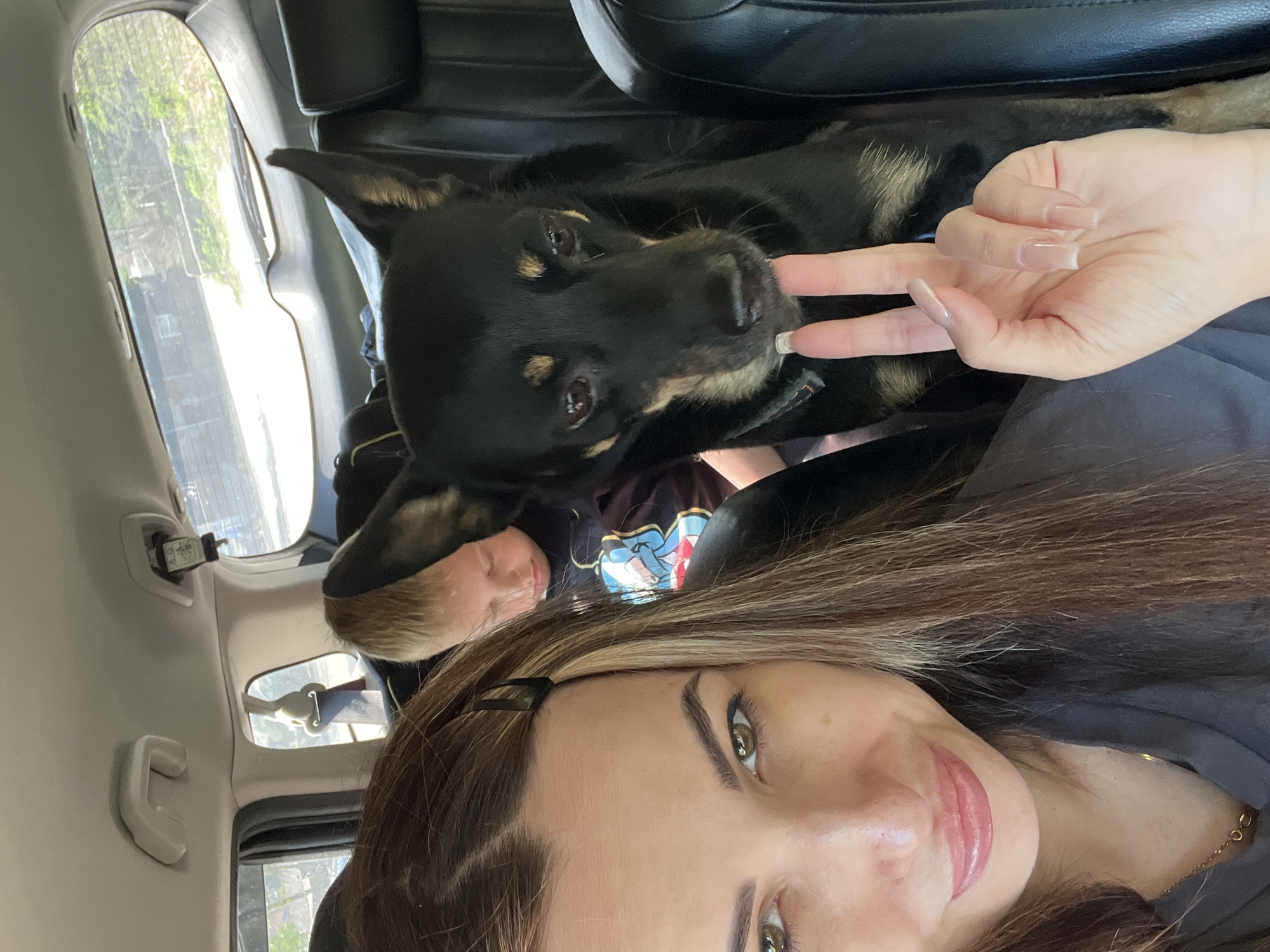 The interior of a vehicle is visible in this selfie-style image. A woman with long brown hair and visible makeup, including eyeliner and lip color, is in the foreground. She has fair skin and a black hair clip is visible in her hair. A hand with long, manicured nails is seen near a black dog that is resting in the back seat of the car. The dog has black fur with brown markings above its eyes and around its muzzle. A person with blonde hair and a t-shirt featuring a graphic is partially visible beneath the dog. The car interior includes a black leather seat and grey panels. Outside the car window, trees and buildings can be seen.