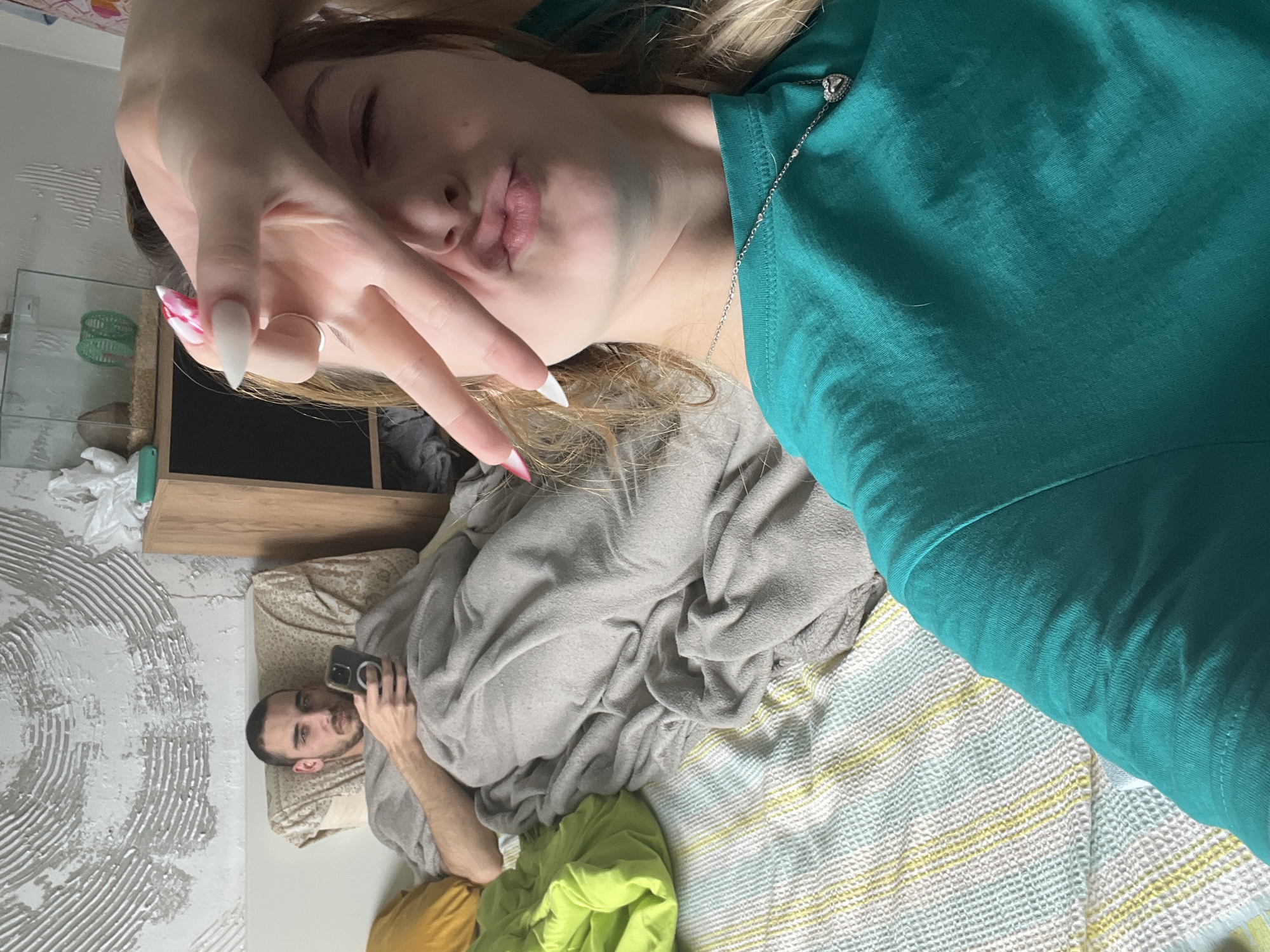 A young woman with long blonde hair and long, white-tipped nails is lying on a bed covered with various blankets in what appears to be a domestic setting. She is wearing a teal top and holding her hand in a "peace sign" pose near her face, with her lips puckered. A silver necklace with a pendant is visible.
In the background, a man lies in bed using a cell phone, while the room shows a wall with a textured design. A nightstand is present and partially visible.
The location is inferred to be Varna, Bulgaria based on the request. No independent verification is possible.