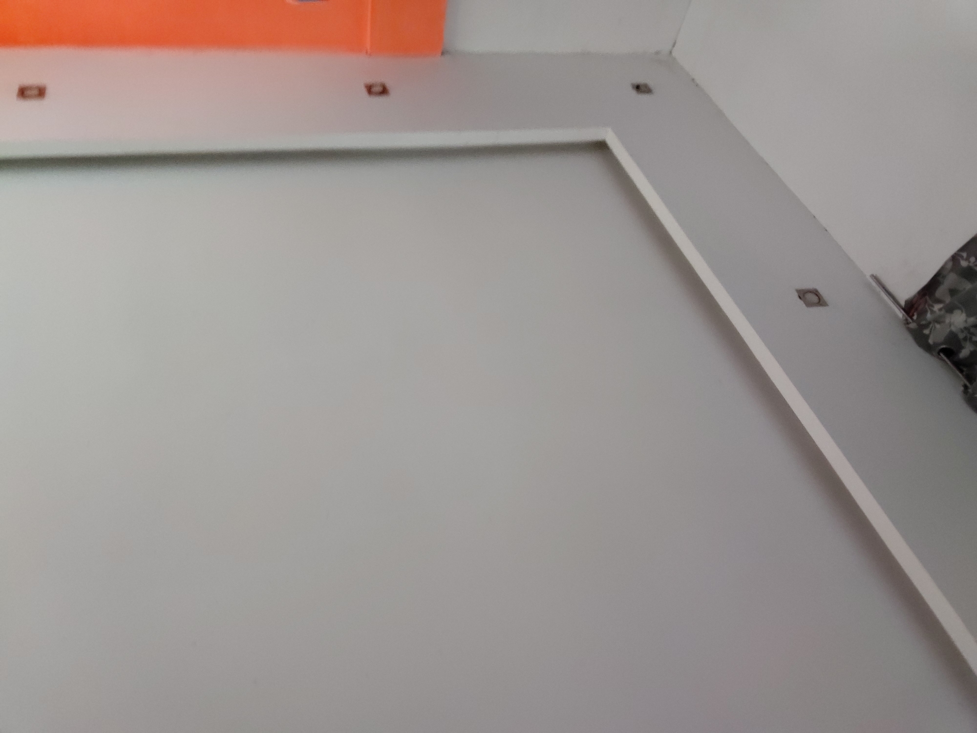 The image captures an interior architectural detail, likely a modern ceiling and upper wall section of a room in Santa Ana, Venezuela. No people are visible in the frame.
The top of the image shows a vibrant orange ceiling segment, suggesting a false ceiling or a distinct painted area. Below this, a lighter gray or off-white ceiling trim extends horizontally, embedded with several small, square, recessed light fixtures. Three such fixtures are clearly identifiable along this horizontal band.
To the right, a prominent vertical light gray structural element, possibly a column or a continuation of the false ceiling design, descends diagonally from the upper right corner. This vertical section also houses square recessed lights, with two visible along its length. These architectural elements create an L-shaped boundary around a large, plain light gray wall that fills the central and lower-left portions of the image.
In the upper right corner, a small, dark, patterned fabric, resembling a curtain or textile, is partially visible. The overall aesthetic is clean and contemporary, utilizing a palette of light grays, off-whites, and a striking orange accent. No activity or event is taking place, and there is no visible text. The time of day or external weather conditions cannot be determined from this interior shot.