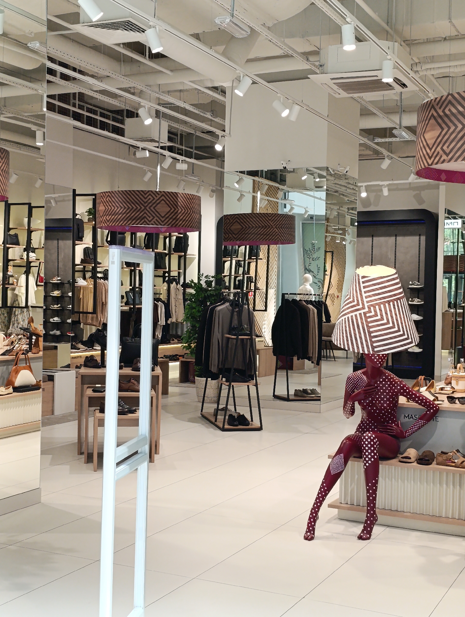 The image depicts the interior of a modern retail store, likely a clothing and accessories boutique, with a focus on shoes. The store is brightly lit with track lighting and large pendant lamps featuring geometric patterns. The walls and ceiling appear to be finished in white and metallic tones, with extensive use of mirrors that create a sense of depth and reflect the store's layout.
The store is organized with tiered shelving units displaying a variety of shoes and handbags. Clothing racks with garments are also visible. The flooring is a light-colored tile.
In the foreground, a mannequin dressed in a form-fitting, dark red suit with white diamond patterns is seated on a bench. The mannequin's pose is relaxed and suggestive, with one hand on its hip and the other touching its chest. It is positioned next to a display of shoes. A large, decorative lampshade with a similar geometric pattern to the ceiling lights is positioned on the mannequin's head.
The scene is orderly and curated, typical of a retail environment designed to showcase merchandise. There are no other people clearly visible in the image besides the mannequin.
The text "MAS**TE" is visible on the bench where the mannequin is seated.
Based on the visual cues and the provided information, this retail store is located in Moscow, Russia.