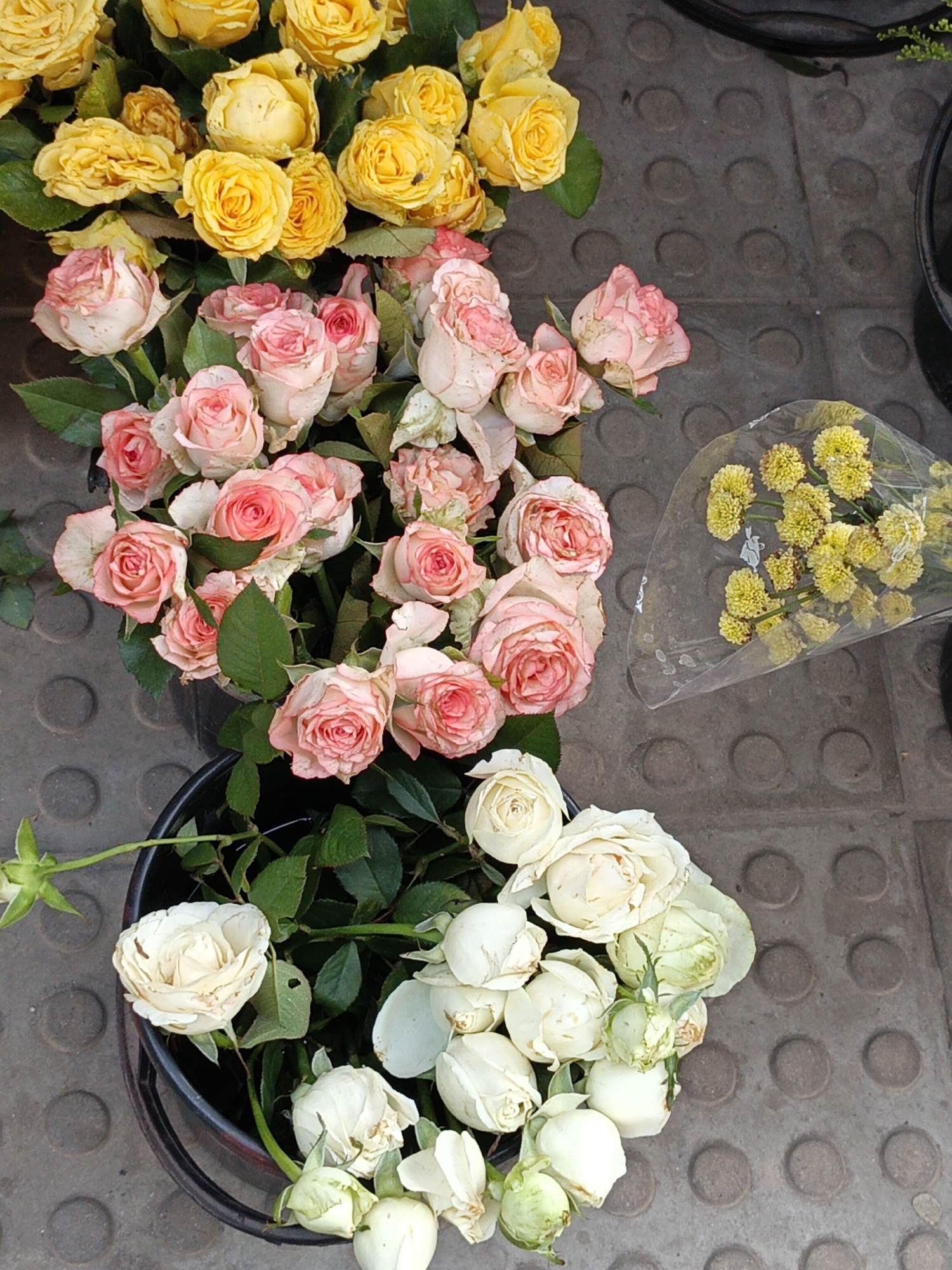 The image shows a collection of flowers, likely for sale, displayed on a gray, patterned surface that appears to be a sidewalk or paved area. There are three buckets containing different colored roses: yellow, pink, and white. A cellophane-wrapped bouquet of small yellow flowers sits to the right of the pink roses. The flowers appear fresh, suggesting they are recently cut or freshly arranged for sale. The image was taken in the city of Dhaka, Bangladesh.