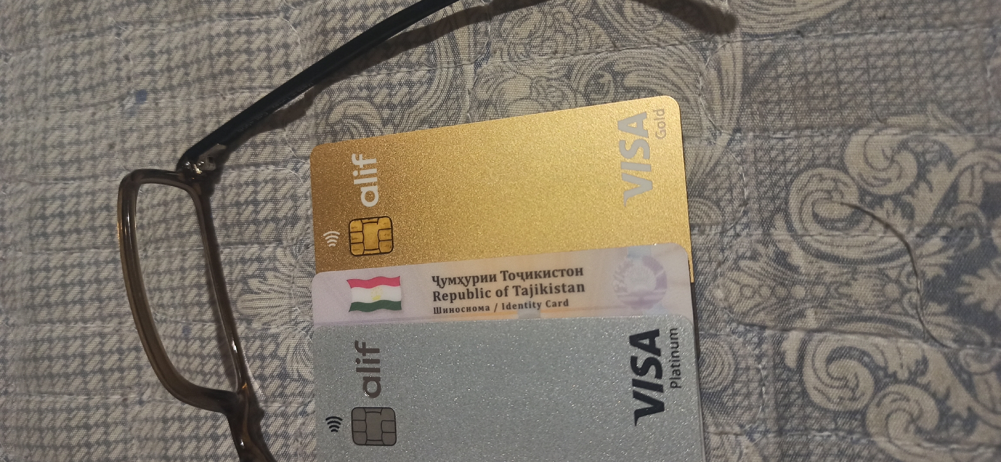 The image displays two Visa cards, a platinum card and a gold card, on a patterned fabric surface, possibly a tablecloth or bedspread. The platinum card is on the left and the gold card is on the right. Both cards have the "alif" logo and a contactless payment symbol. The platinum card has "VISA Platinum" printed on it, while the gold card has "VISA Gold" printed on it. The fabric has a detailed, swirling pattern in shades of blue and white. A pair of eyeglasses, with dark brown frames, is positioned at the top of the image, partially obscuring the cards. A thin black wire, possibly from the eyeglasses, also runs across the fabric. A small portion of the Republic of Tajikistan flag is visible between the two cards, along with text in Cyrillic and English, indicating "Republic of Tajikistan" and "Identity Card". The scene is an indoor still life.