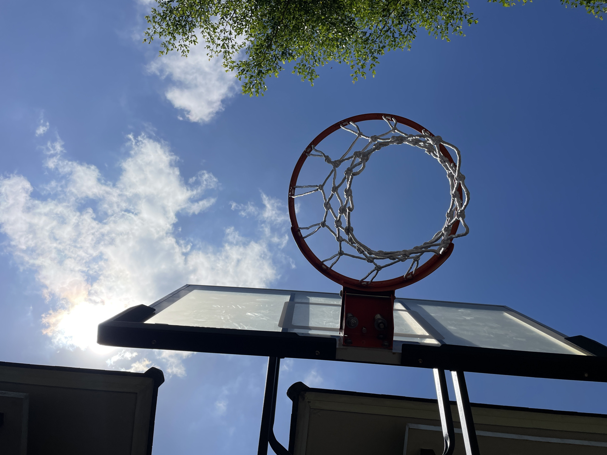 A low-angle view captures a basketball hoop with a white net against a bright blue sky with scattered clouds and green foliage at the top. The hoop is orange and mounted on a clear backboard with a black frame, which is attached to a support structure. The support structure is placed atop a building. The low-angle viewpoint suggests the photo was taken from ground level. Sun glare is visible through the backboard. The location is specified as Kuta Selatan, Indonesia.