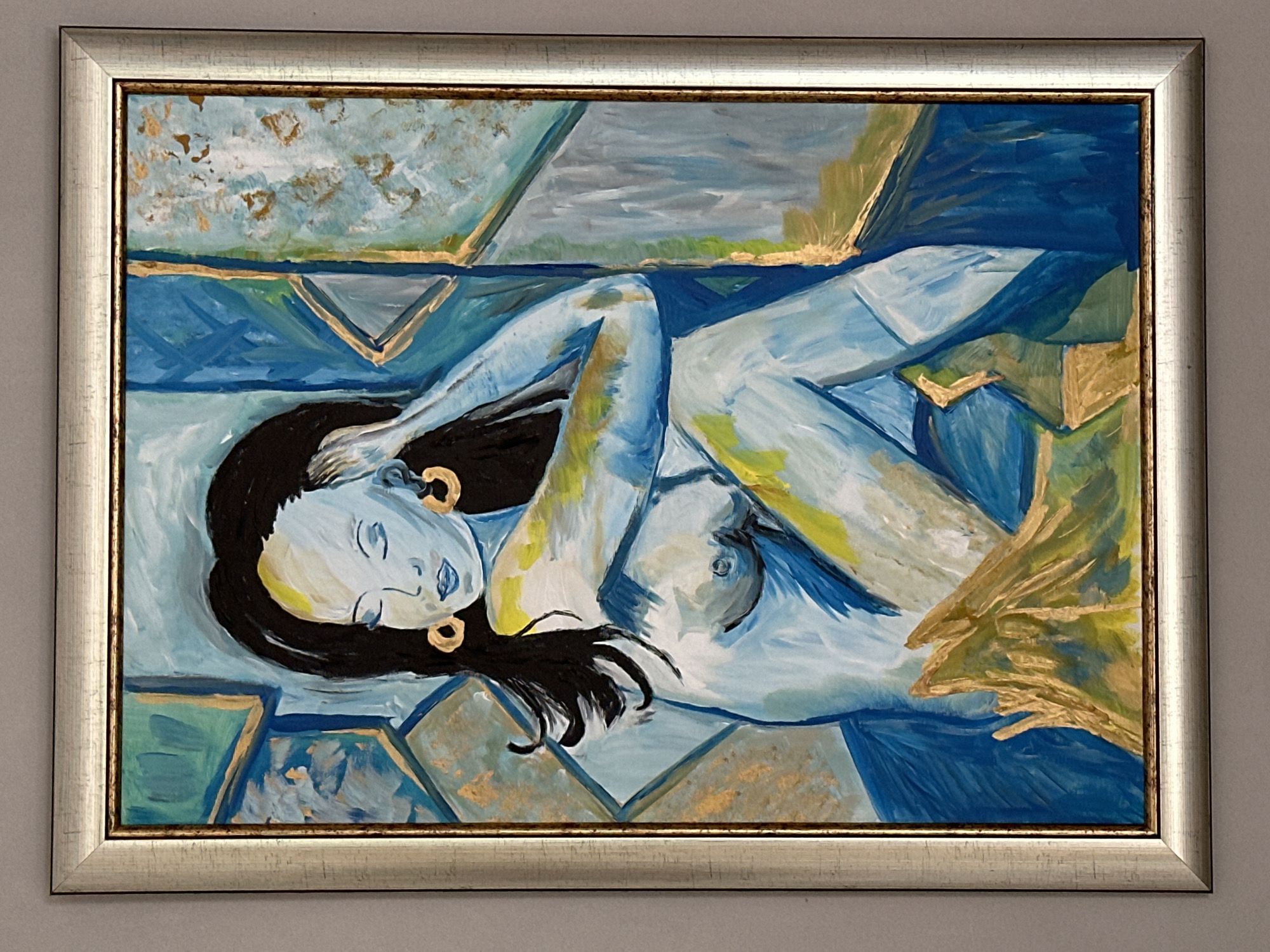 The image is a framed painting of a nude woman with blue skin, dark hair, and closed eyes. She is sitting with her legs crossed and her left arm resting on her knee, while her right hand is touching her hair. She is wearing large gold hoop earrings. The background is abstract and features shades of blue, green, and gold, with geometric shapes. The painting is framed in a metallic silver frame. There is no indication in the image or its context that this painting is located in the city of Glavatartsi, Bulgaria.