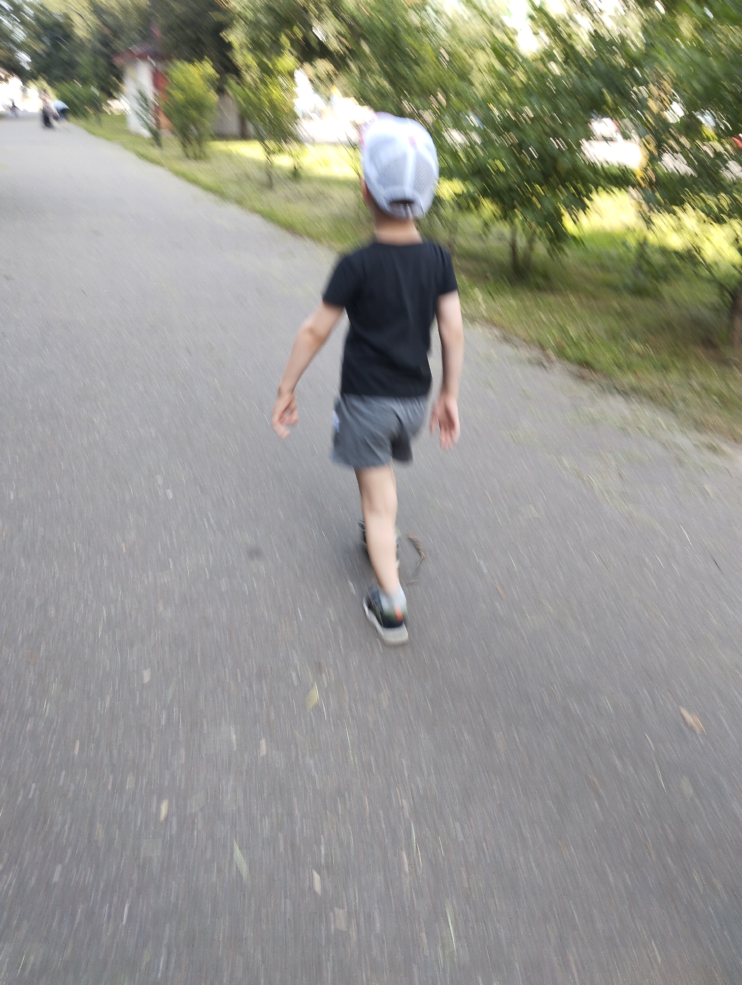 The image captures a person walking away from the camera on a paved path, likely in Kingisepp, Russia, given the context. The individual is wearing a black t-shirt, grey shorts, a white and grey baseball cap, and dark sneakers with white soles and an orange accent on the heel. The path appears to be lined with trees and grass on the right side. The image has a motion blur effect, indicating movement. In the background, on the left side of the path, there are hints of buildings and other people walking, though they are indistinct due to the blur. The overall scene suggests a casual outdoor activity in a park or recreational area.