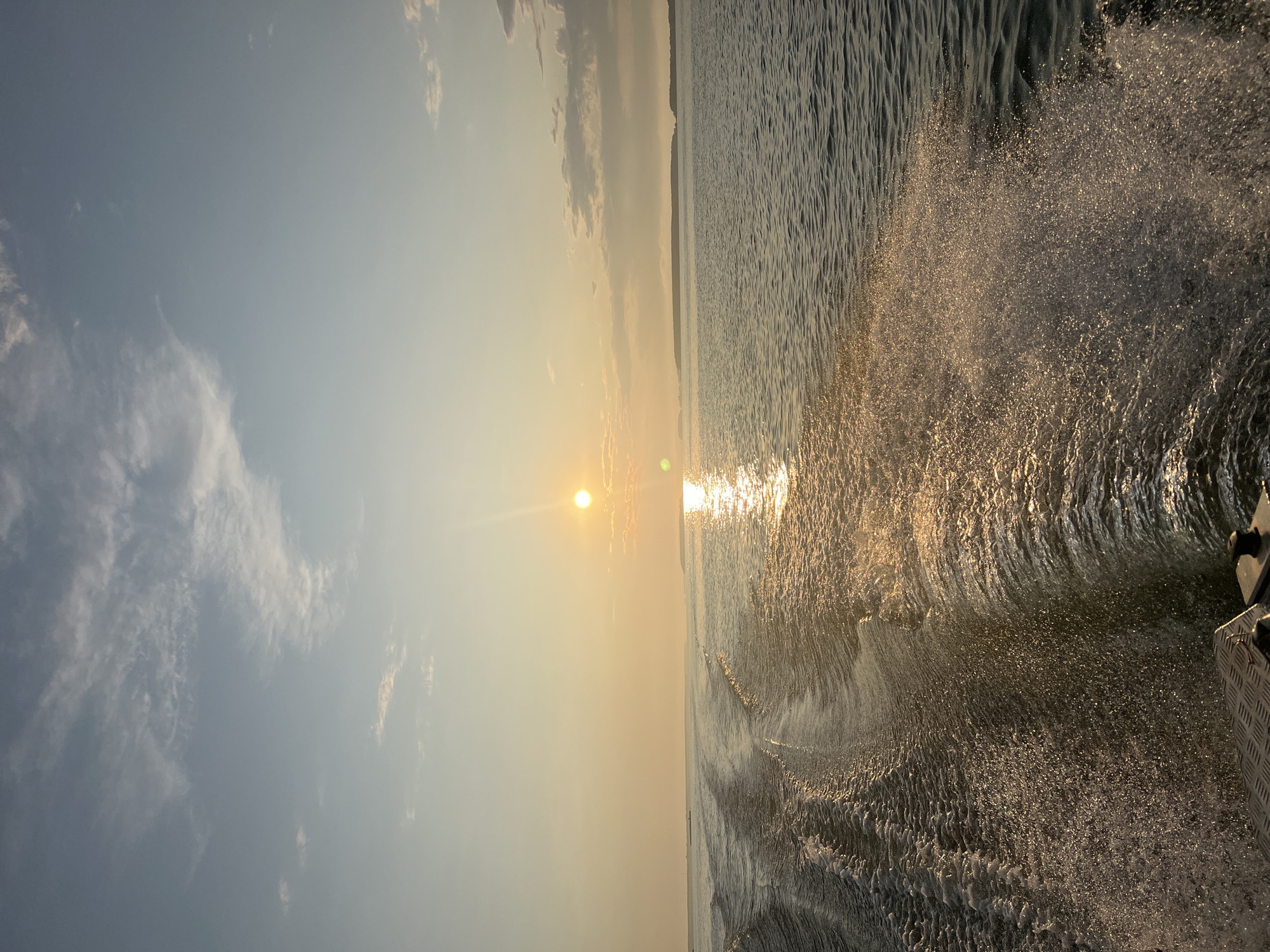 This is a wide-angle, eye-level shot of a wake behind a boat moving across a body of water. The sun is setting, casting a warm, golden light across the scene, with the sun itself visible as a bright orb in the upper center of the frame, reflecting a shimmering path on the water. Wispy clouds are scattered across the pale blue sky. In the distance, a landmass with gentle hills is visible on the horizon. The foreground shows the textured metal of the boat. The water is a deep blue-grey, with the wake creating foamy white patterns and splashes. The image appears to be taken from the stern of the boat. The location is specified as Novopichugovo, Russia.