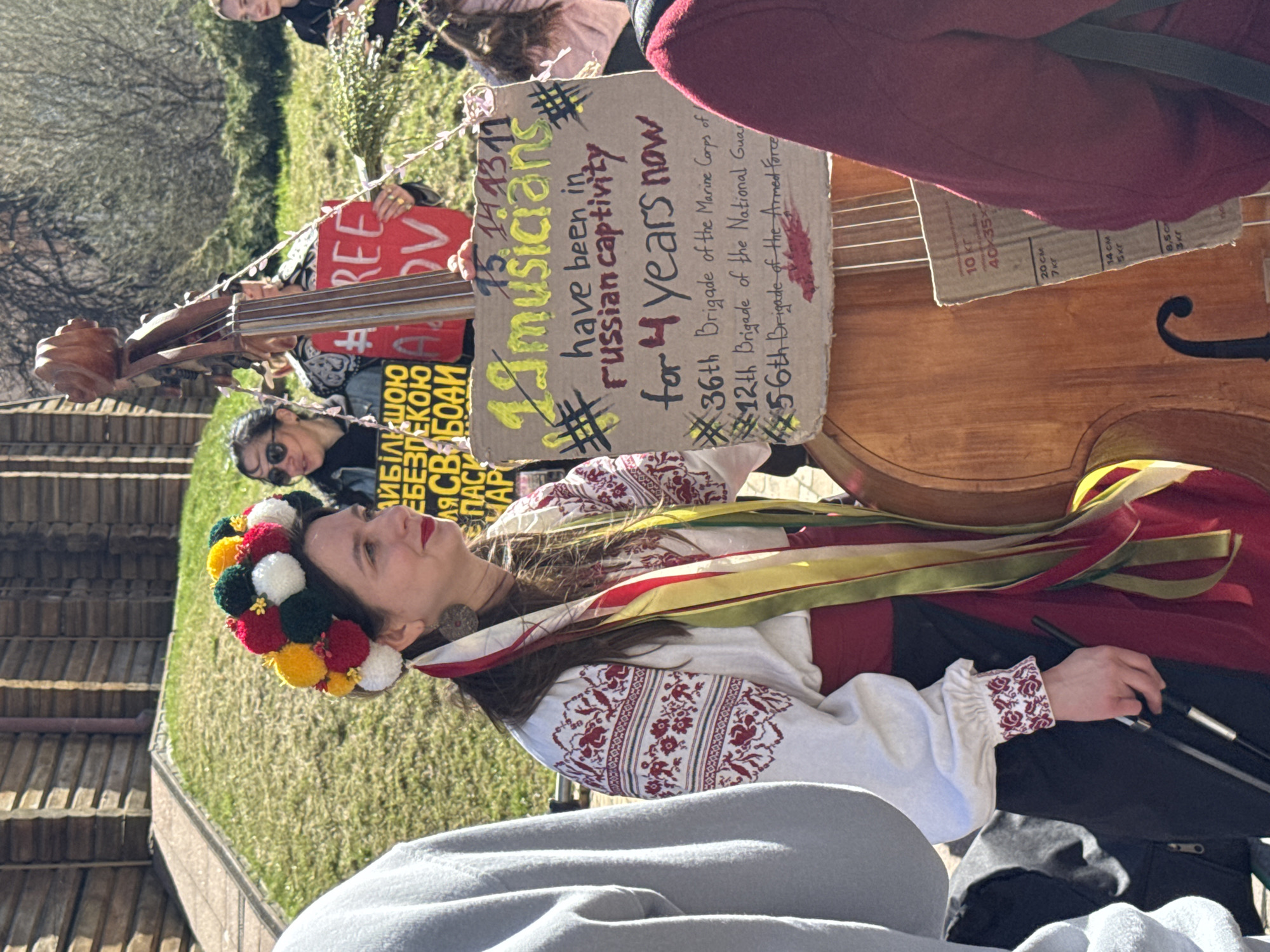 The image shows a demonstration in Kyiv, Ukraine. A woman in traditional Ukrainian dress stands playing a double bass. Attached to the bass are two signs. One reads "FREE ADV" and another reads "#13, 14, 13, 11, 19 musicians have been in Russian captivity for 4 years now, #36th Brigade of the Marine Corps of, #12th Brigade of the National Guard, #56th Brigade of the Armed Force". Another sign reads "АЙБІЛЬШОЮ ЕБЕЗПЕКОЮ ДЛЯ СВОБОДИ СПАСИ АР" (More danger to freedom save AP). People are standing around the woman. The environment includes buildings, trees, and grass.