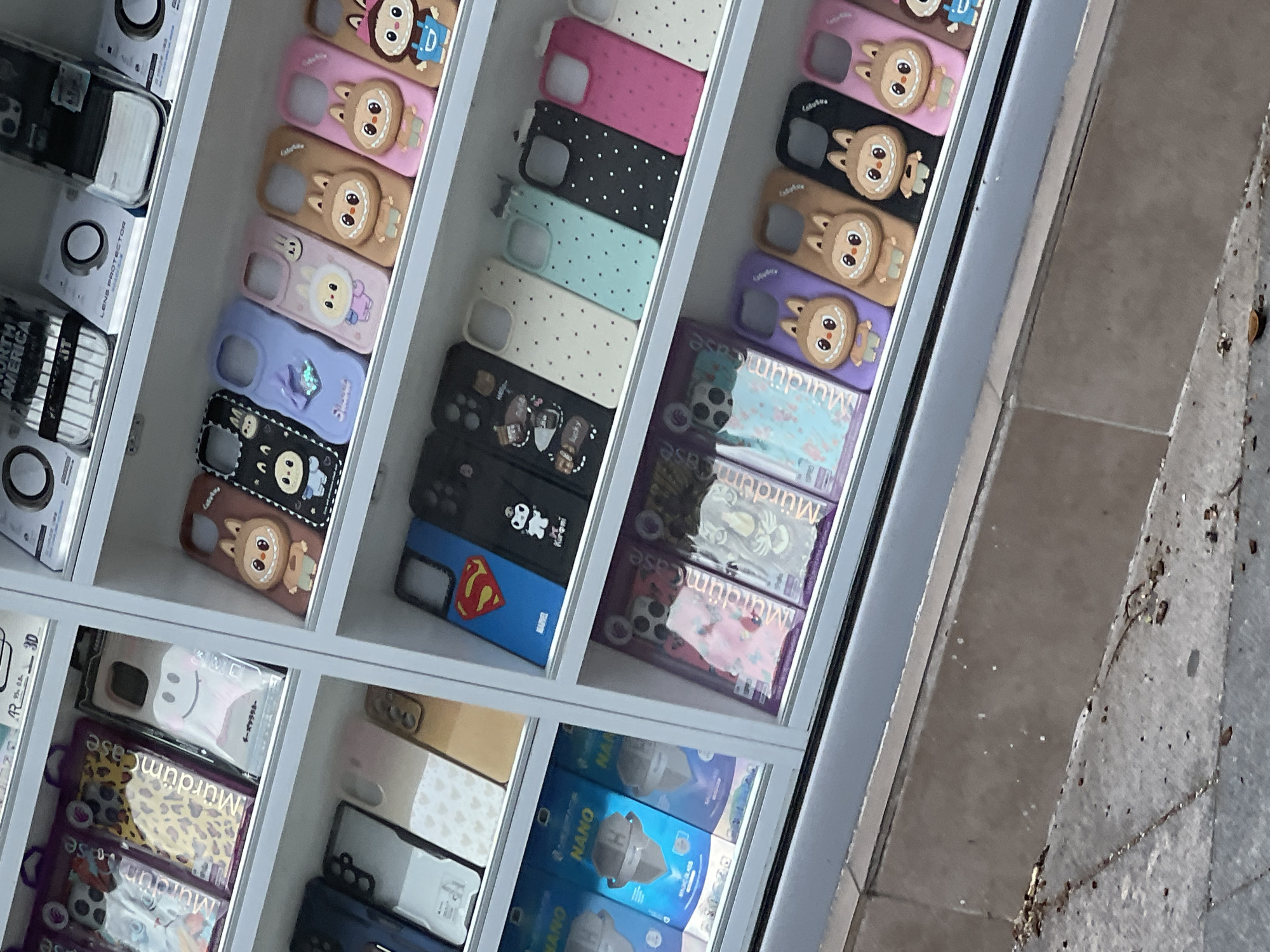 Here is a concise description of the image:
The image shows a display case, likely outside a store, filled with various phone cases. The phone cases have different colors, patterns, and designs. Some have cartoon characters, while others have simple geometric designs. There are also some packages of phone protectors. The display case is located near a sidewalk, as evidenced by the concrete visible on the right side of the image. The text on the packages and phone cases suggests the location might be in a Bulgarian-speaking area, such as Plovdiv, Bulgaria, given the presence of the word "Murdüm."