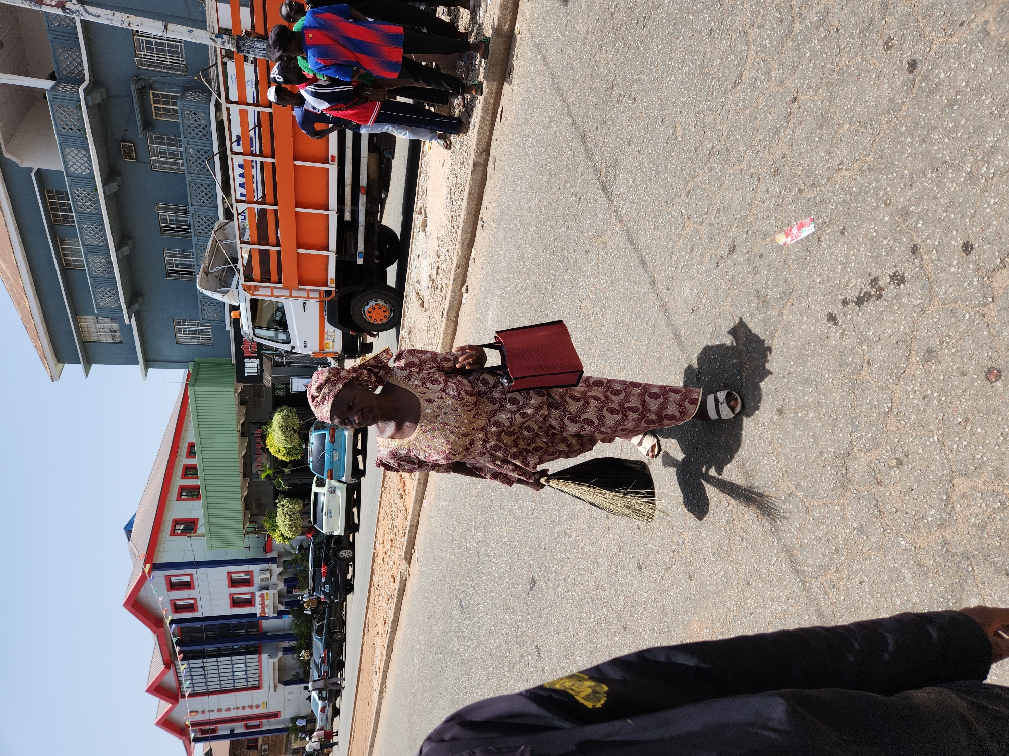 A medium shot depicts an older woman walking on a paved street in Unknown, Unknown. She wears a patterned dress and headwrap, white sandals, and carries a red bag in her left hand and a broom in her right. A truck is parked on the side of the street and people are standing beside it. Buildings are also on the side of the street, as well as several parked cars. A piece of trash sits on the road. The lower portion of a person in a black jacket is shown in the foreground.