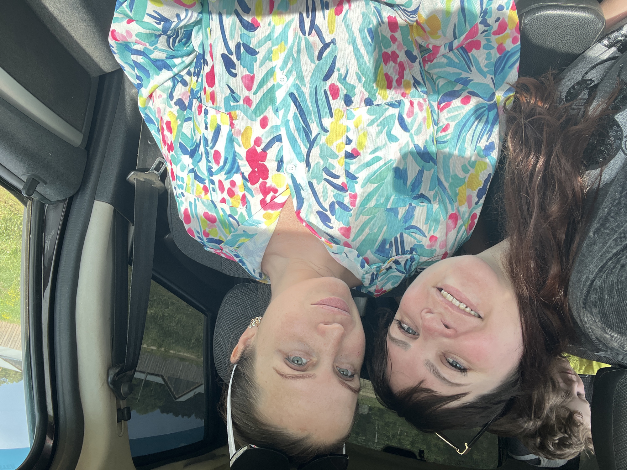 Two women are posing for a selfie inside a vehicle. The woman on the left has light skin, blue eyes, and is wearing a floral-patterned blouse. Sunglasses are placed on her head. The woman on the right has darker hair and is wearing a gray shirt. They are both smiling. The vehicle's interior is visible, including the seats, windows, and seatbelts. The outside environment appears to have green foliage and a building. The image is taken at an odd angle. There is also a young child visible behind the woman in the gray shirt. The location is not mentioned.