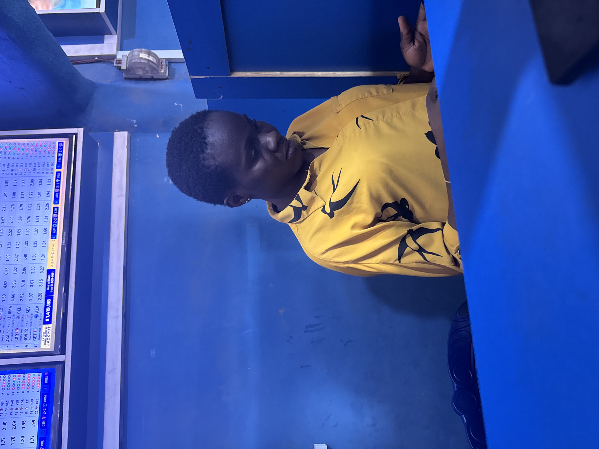A person is sitting at a blue counter in a room with blue walls. The room is dimly lit. A betting terminal displaying odds and sporting event details is visible. The odds include teams like GEN, SAS, CAG, VER, TOR, LEC, PAR, and CRE. The display also indicates "KING MAKER JACKPOT" with a value of "#1,419,138" and "Won in Rivers." The location of this establishment is likely in Nkpor, Nigeria. The person is wearing a yellow shirt with black designs. Their hand rests on the counter.