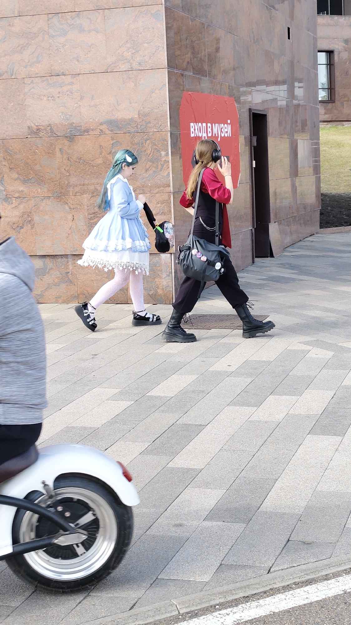 The image depicts two individuals walking along a paved walkway in front of a large, reddish-brown tiled building. A sign with text in Russian reading "ВХОД В МУЗЕЙ" (Entrance to the Museum) is visible on the building.
The first individual is dressed in a blue lolita-style dress with white lace and white stockings. They have blue hair and are carrying a black bag. The second individual is wearing a red and black outfit, including a red shirt, black pants, and black boots. They have long brown hair and are wearing headphones. They have a gray bag with buttons slung over their shoulder.
A person on a small white motorized scooter partially obstructs the view on the left side of the image. The ground is paved with gray rectangular stones, and a white line is visible along the edge of the walkway. In the background, there is some greenery and part of another building.