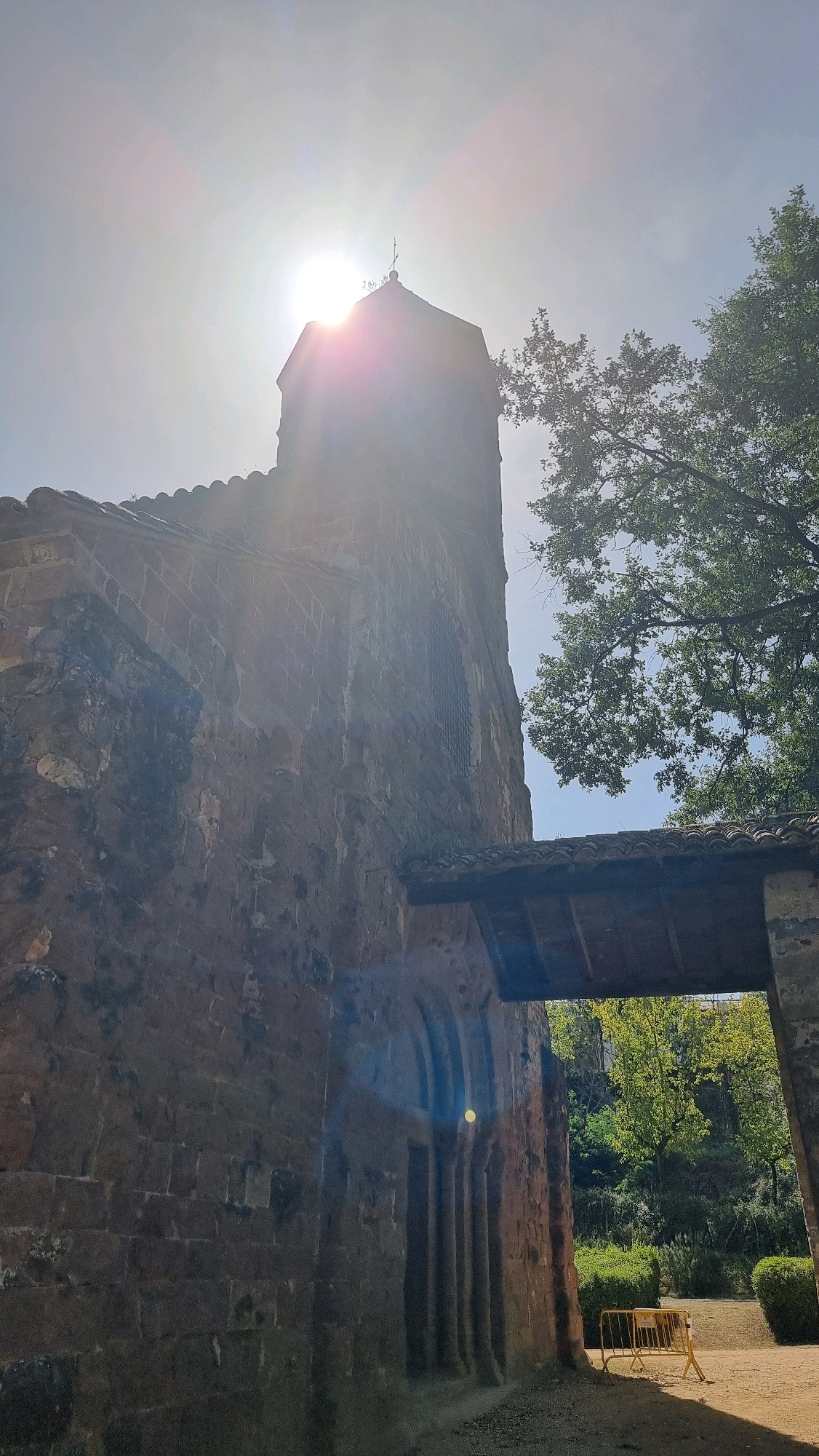 This is a low-angle, outdoor, medium shot of a stone building, possibly a church or ancient structure, with a prominent tower. The sun is very bright and directly overhead, creating a lens flare and washing out the sky. The building's stone walls are rough and appear weathered. A wooden covered walkway extends from the building. To the right of the building, a large tree with dense green foliage is visible, its branches reaching towards the structure. In the background, there is more greenery and a small yellow barrier fence. The lighting suggests it is daytime, possibly late morning or early afternoon. The overall impression is one of historical architecture set against a natural backdrop. The location is identified as Sant Joan les Fonts, Spain.