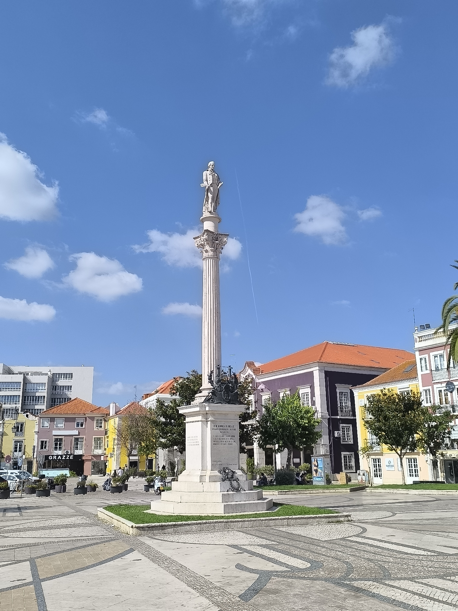 This is a vertical, eye-level shot of a public square in Setúbal, Portugal, under a bright blue sky dotted with fluffy white clouds. The sun is shining, suggesting it's daytime, likely late morning or early afternoon given the shadows.
The central subject is a tall, ornate white monument topped with a statue of a standing man. The monument has a detailed column and a pedestal with inscriptions and further sculptures. The monument stands in the foreground, on the right side of the frame, dominating the center of the square.
The square itself is paved with intricate patterns of stone and tiles. Green grass surrounds the base of the monument. In the background, a row of colorful buildings lines the square. These buildings feature various architectural styles and colors, including yellow, purple, and orange roofs. Some of these buildings appear to be commercial establishments, with visible signage. One sign reads "GRAZZE". Another building has a sign that says "SABSEG".
Several people are visible in the square, either walking or sitting. Some are near the monument, while others are near the buildings. A few scooters or small motorcycles are parked in front of some of the businesses, and there are also planters with greenery placed around the square. The overall atmosphere is one of a peaceful, everyday scene in a European city. The presence of a contrail in the sky suggests air traffic.