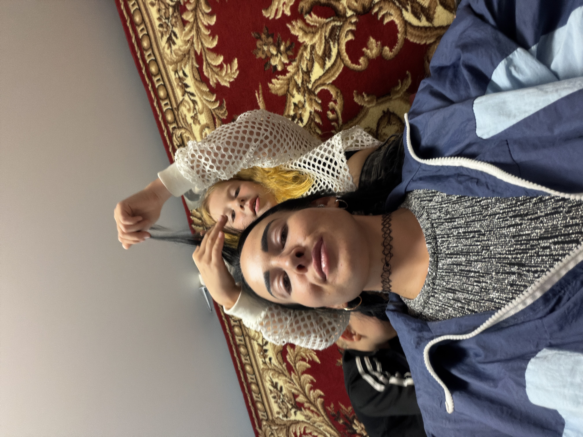 A low-angle, head-on, waist-up shot captures three individuals indoors, positioned in front of a red and gold patterned rug that covers the wall and floor. In the foreground, a woman with dark hair and a choker necklace smiles at the camera, wearing a gray, ribbed shirt and a blue and light blue zippered jacket. Her hands are clasped in front of her. Behind her, a person with dark hair is partially visible to the left, wearing a black and white jacket. To the right and slightly behind the woman, a young girl with blonde hair and a white mesh top is seen from the shoulders up, reaching to touch the woman's hair. The lighting suggests an indoor setting, possibly a home in Бронницы, Russia.