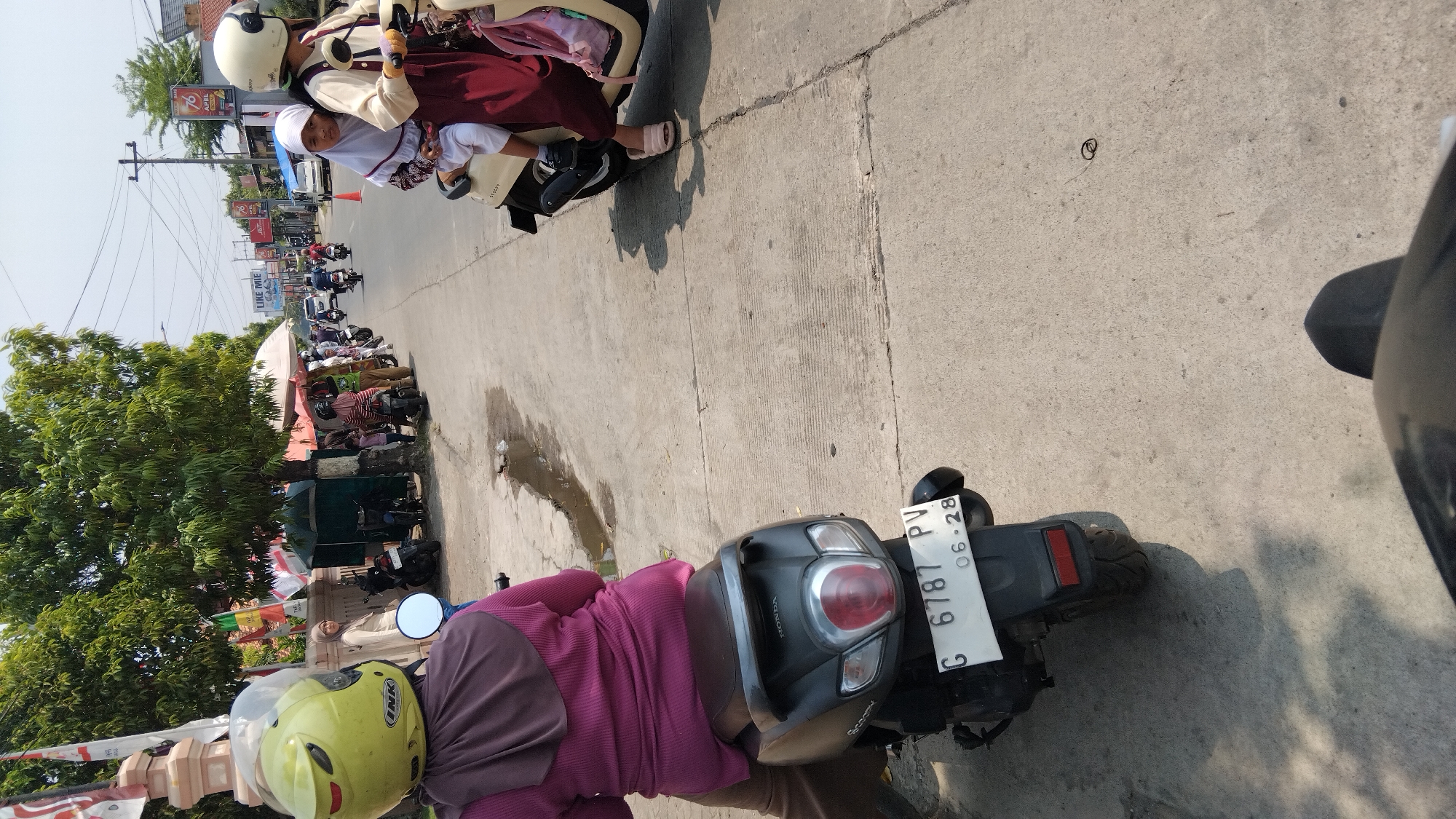 In Watesalit, Indonesia, a street scene unfolds under a bright sky. A person, wearing a purple long-sleeved shirt and a brown hijab, sits on a black Honda scooter. The scooter's license plate reads "G 6787 PV 06-28." To the right, another scooter carries two individuals; one wearing a white hijab and a white and maroon outfit, and another in a white hijab and a maroon dress. The background reveals a road lined with trees, shops, and several other motorcycles. A woman in a light-colored hijab stands near a wall with flags. The ground is paved with concrete, showing some water stains and cracks.