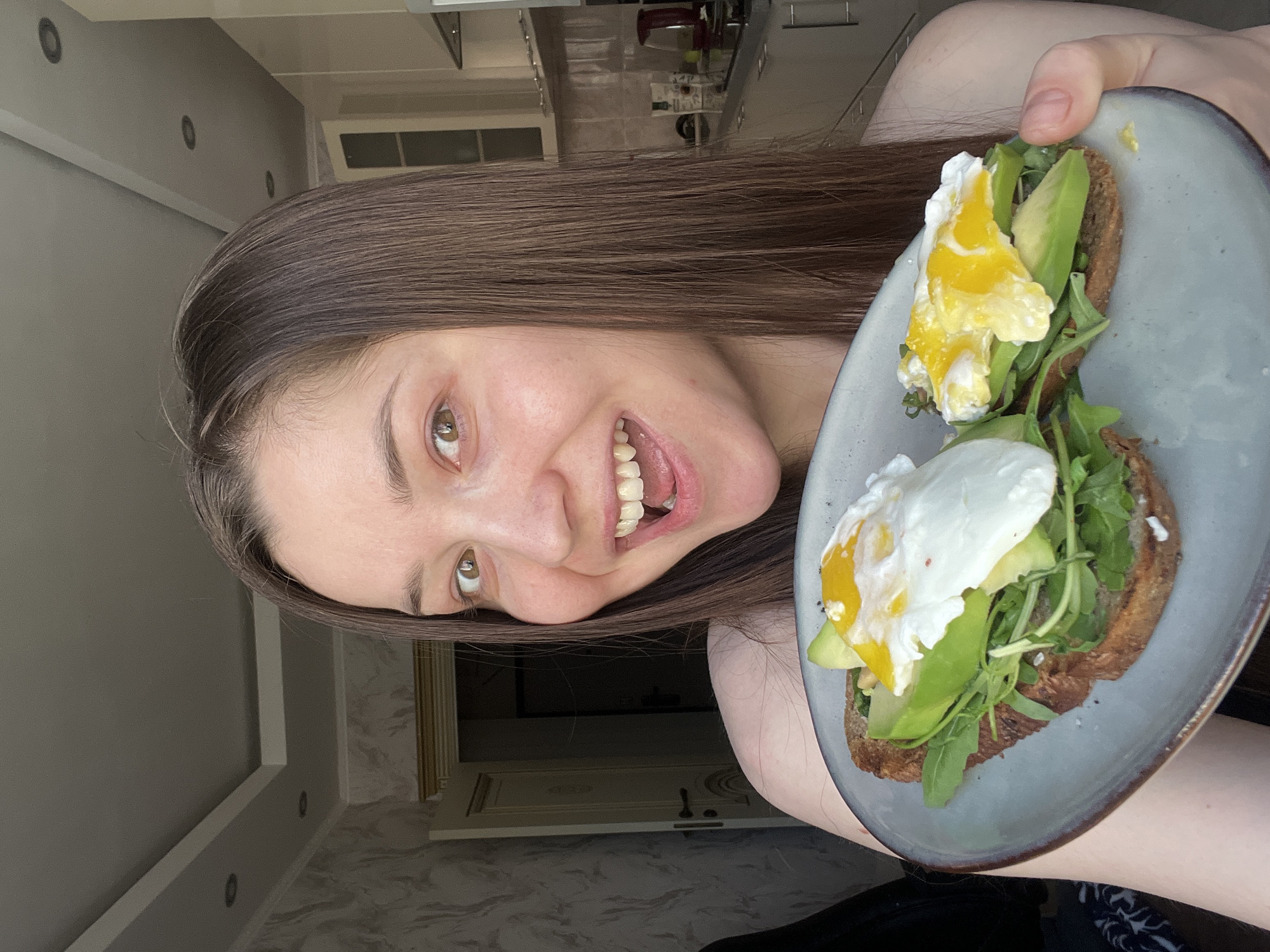 A woman with long brown hair is holding a plate with two pieces of toast topped with avocado, arugula, and a fried egg. She is smiling at the camera. The background appears to be a kitchen, with cabinets and a countertop visible. The location cannot be determined as Khimki, Russia.