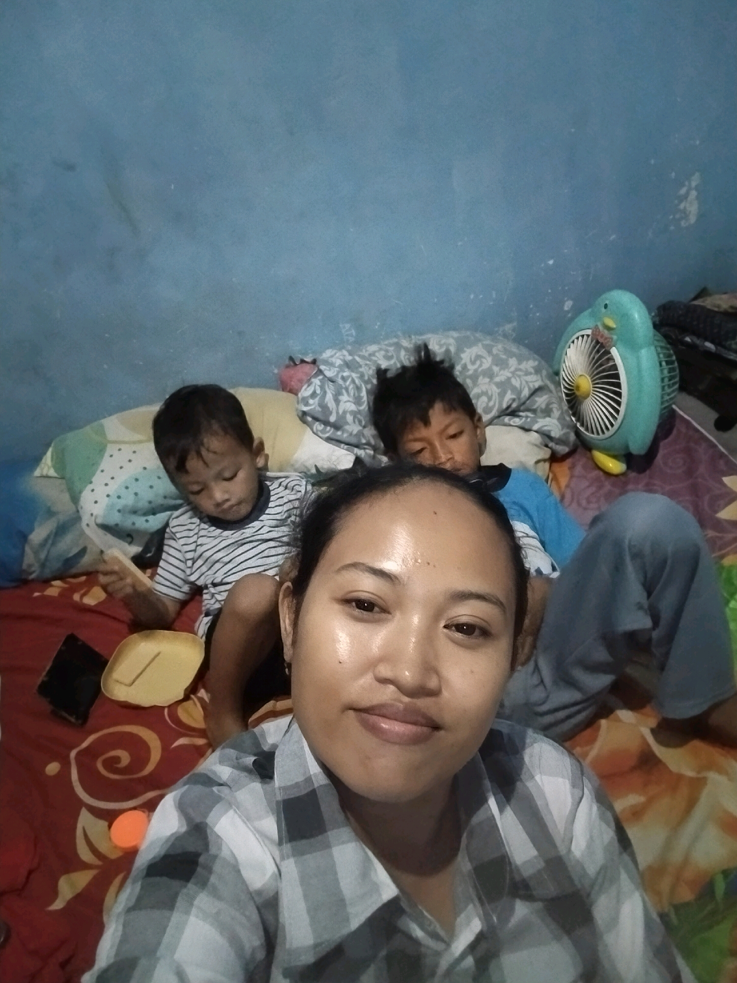 The image captures a selfie taken by a woman in a bedroom in Beloh, Indonesia. The woman is in the foreground, smiling and looking at the camera. Behind her, two young boys are lying on a bed. The boy on the left is wearing a striped shirt and holding a yellow object. The boy on the right is wearing a blue shirt and looking towards the camera. The room is dimly lit, with blue walls and a patterned bedspread. A small turquoise fan shaped like a duck is visible on the right side of the image. There is a dark object, possibly a phone, on the bed to the left of the woman. The overall impression is one of a casual, intimate family moment.