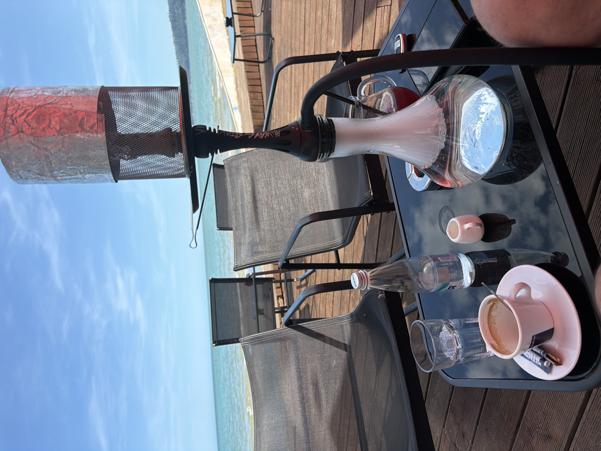 A hookah sits on a table by the sea in Varna, Bulgaria. The scene depicts a waterfront cafe with dark chairs and a wooden deck. The hookah is made of glass and metal, and smoke is visible. On the table, there is a cup of coffee, a glass of water, and a bottle of water. The sea and the sky are visible in the background.