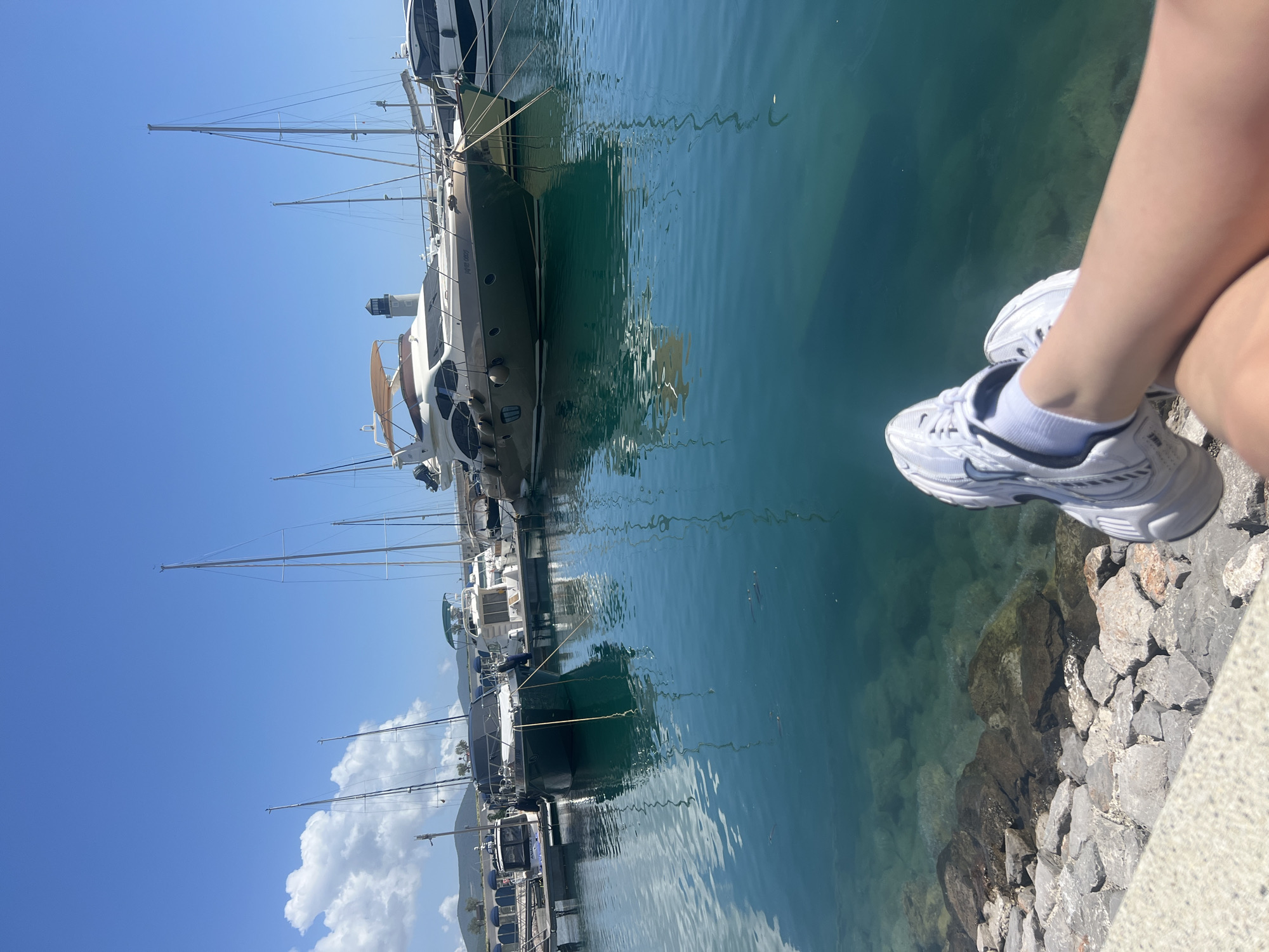 A person's legs, wearing white sneakers and socks, are positioned over a rocky edge and the water, likely a harbor, with docked yachts in the background, which is reflected in the clear water. The sky is blue with some clouds. The image was likely taken in Radovići, Montenegro, based on the presence of the yachts and the distinctive coastal landscape.