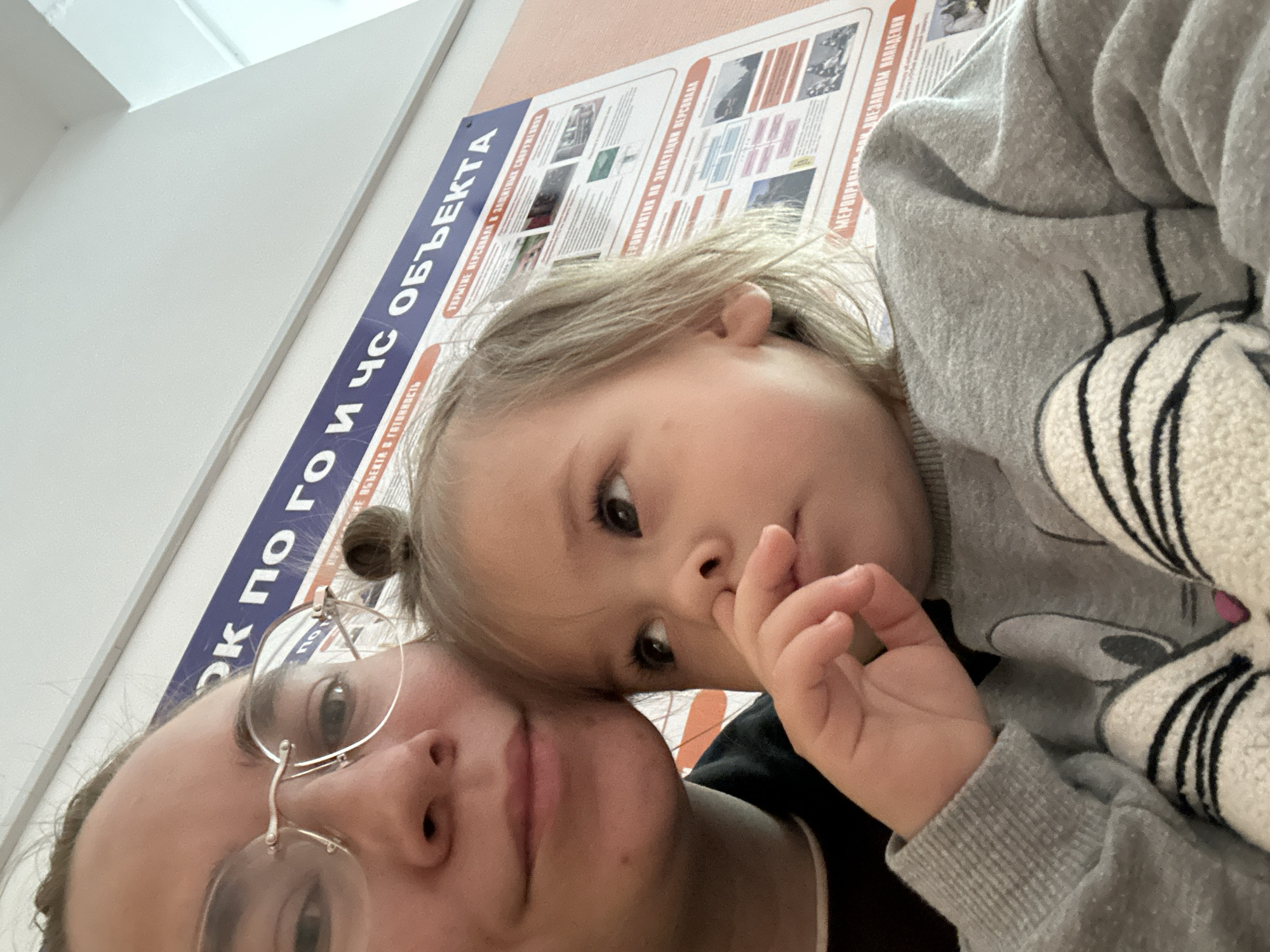 Here is a detailed description of the image:
**General Description:**
The image is a selfie taken indoors, likely by an adult female wearing glasses, with a young child (likely a toddler). The photo is taken at an angle, with the top of the image tilted to the left.
**People:**
* **Adult Female:** She is wearing glasses and has a portion of her face visible, including her eyes, nose, and mouth. She appears to be the one taking the selfie.
* **Young Child:** The child is close to the adult, possibly being held or leaning against her. The child has fair skin, light hair, and is wearing a grey sweater with a black and white cartoon-like figure on it.
**Objects and Environment:**
* **Wall:** A wall is visible behind the subjects.
* **Poster/Sign:** Affixed to the wall is a poster or sign with text in what is identified as Russian, partially obscured by the subjects.
* **Clothing:** The child is wearing a grey sweater. The adult is wearing a dark top.
**Text on the Poster:**
The text on the poster appears to say: "ОК ПО ГО И ЧС ОБЪЕКТА" and includes words such as "УКРЫТИЕ ПЕРСОНАЛА" and "ЭВАКУАЦИИ ПЕРСОНАЛА".
**Actions and Interactions:**
* The adult is holding the camera and taking the picture.
* The child is close to the adult, possibly interacting by leaning against or being held. The child has one finger in its mouth.
**Location Context (Chaikovsky, Russia):**
* The Russian text on the poster suggests the location is in a Russian-speaking area, most likely Russia. The phrase "ОК ПО ГО И ЧС ОБЪЕКТА" suggests a sign related to civil defense or emergency response protocols for a facility or organization.